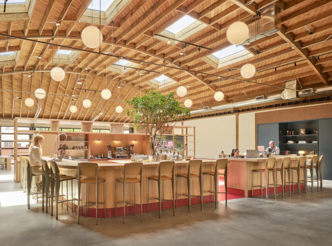 Bright, spacious bar area featuring a soaring, wooden-beamed ceiling with circular pendant lights. Light spills in through several skylights. A central bar made of warm wood is surrounded by modern bar stools, complemented by lush greenery, creating an inviting social atmosphere.
