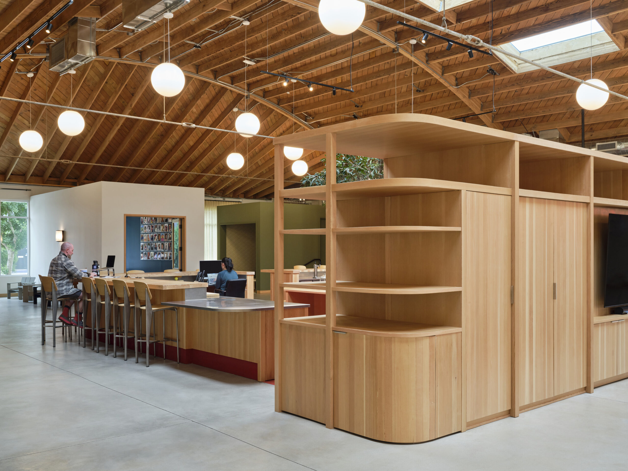 Warm wood tones define this contemporary open-plan workspace, featuring curved shelving that adds a soft touch. Pendant lights enhance the airy atmosphere, while large windows bring in natural light. A mix of seating areas and workstations promotes collaboration, creating an inviting and functional environment for productivity.