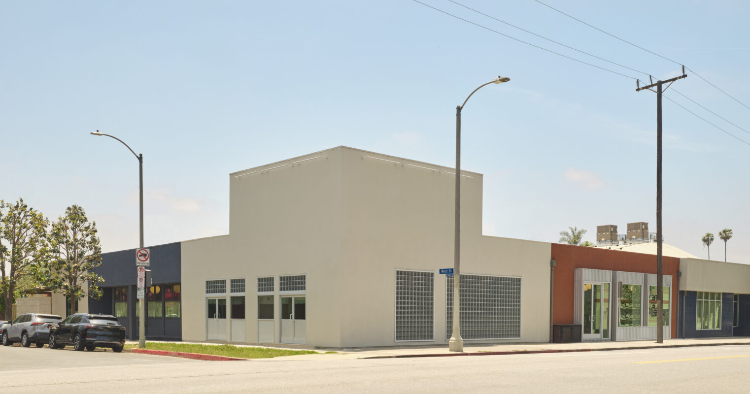 Contemporary commercial building featuring a mix of colorful facades, large glass windows, and geometric shapes. The structure includes a prominent white section, contrasting with adjacent hues of blue and orange. Landscaping elements soften the urban setting, enhancing the overall aesthetic appeal and functionality of the design.