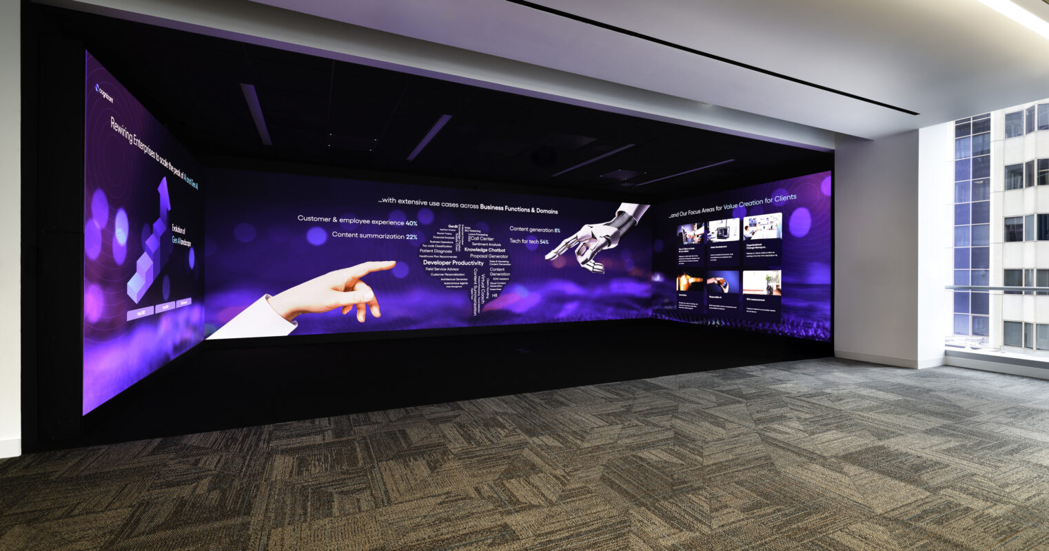 A modern, spacious interior with a large digital wall displaying interactive content in shades of purple. The room features textured gray carpeting and sleek black ceiling lines, creating a professional environment that encourages collaboration and innovation. The lighting enhances the technological focus of the space.