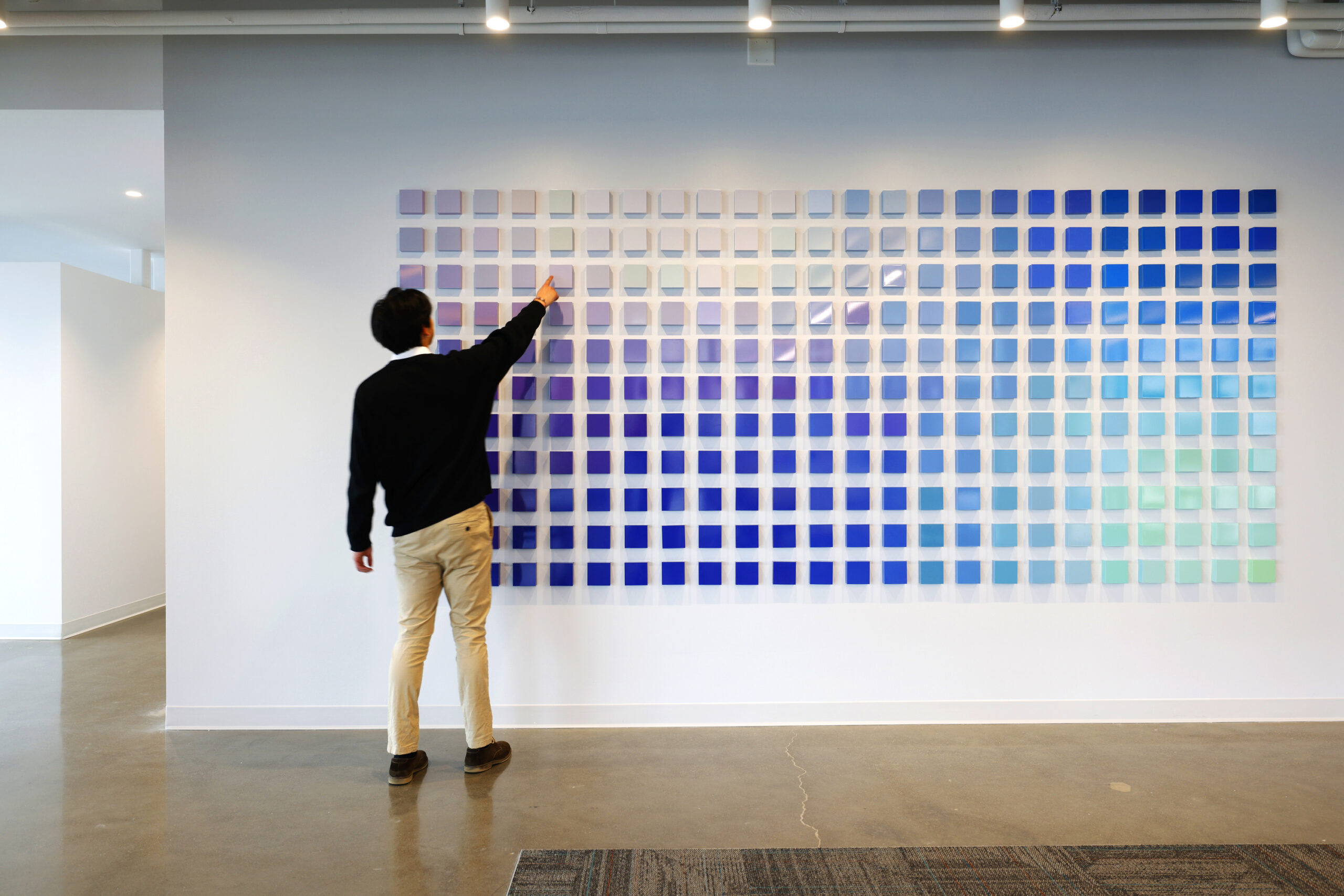 A designer evaluates a vibrant wall installation featuring a grid of 100 square color swatches in varying shades of blue, green, and gray. The artwork serves as a dynamic focal point, contributing to the modern, open-concept office environment with polished concrete flooring and minimalist decor.