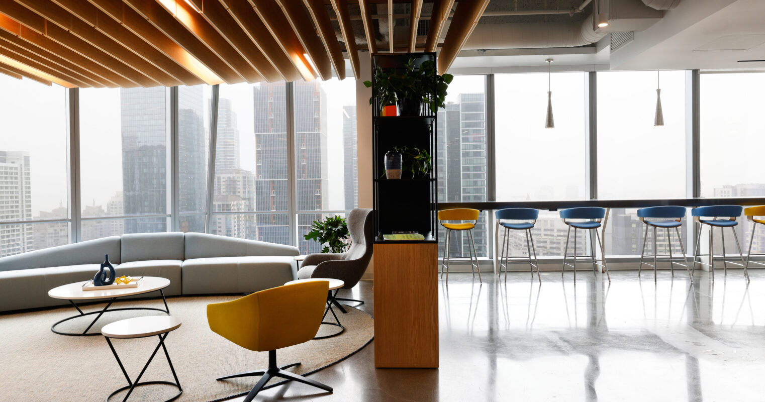Contemporary office lounge featuring a modern aesthetic. A curved gray sofa and a vibrant yellow chair offer comfortable seating. Natural light floods the space through large windows, accentuated by a wooden slatted ceiling. Sleek bar stools line a high table, creating an inviting social atmosphere.