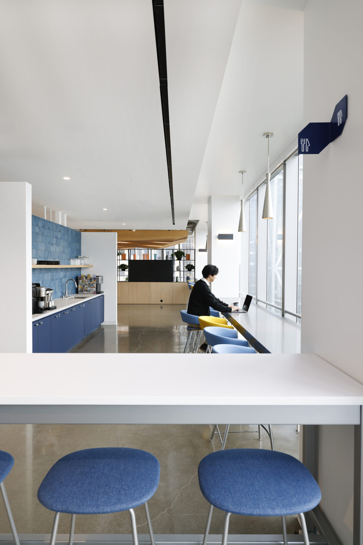 Modern office kitchen featuring a minimalist design with a sleek blue cabinetry and polished concrete flooring. A long white counter with blue stools offers seating, while large windows provide abundant natural light. A person is seen working on a laptop, highlighting the functional and social aspects of the space.