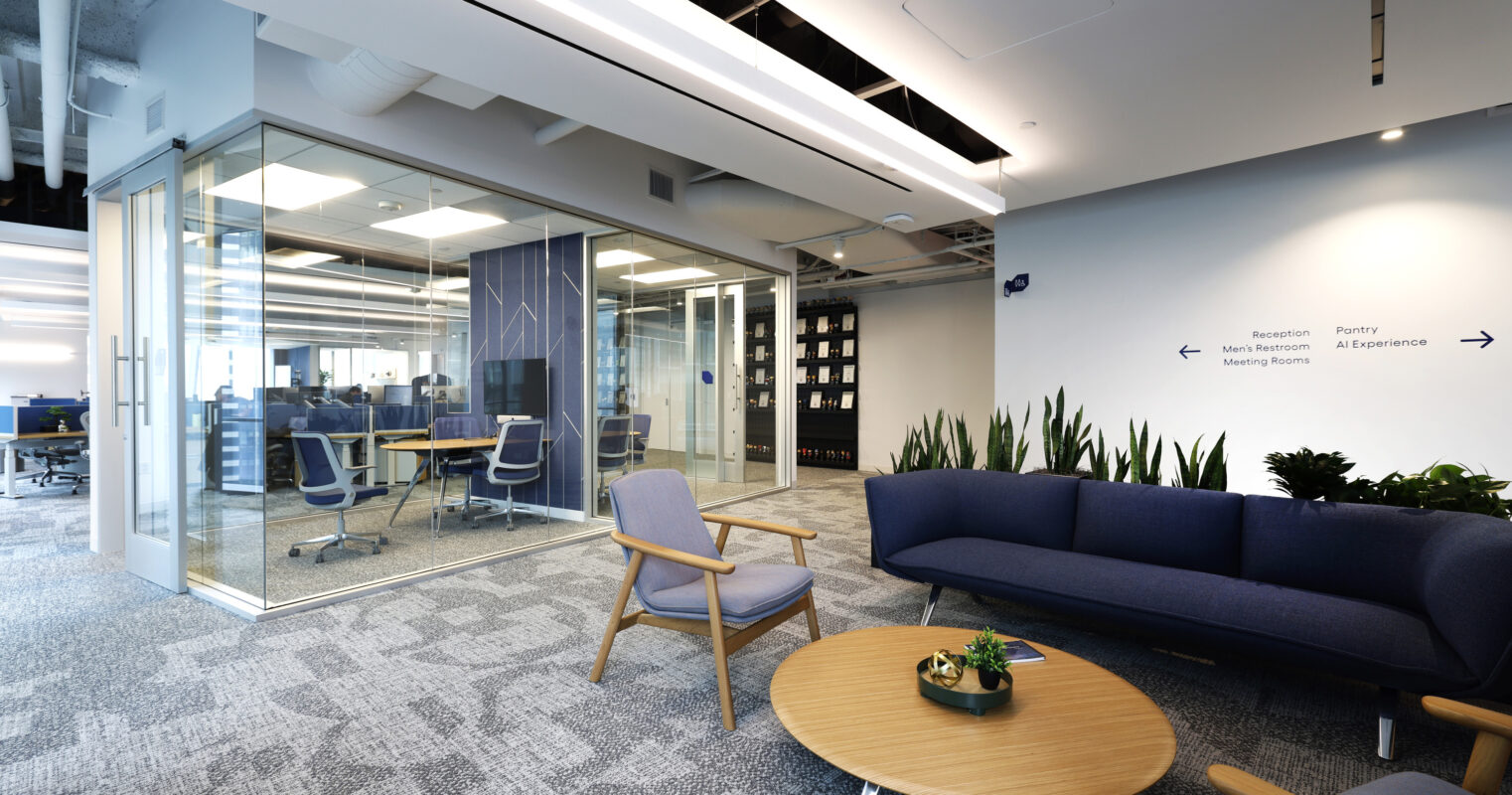 A modern office lounge features a blue sofa paired with a round wooden coffee table. Elegant chairs and potted plants enhance the inviting atmosphere. Glass walls enclose meeting rooms in the background, while the textured carpet adds depth to the contemporary design, promoting collaboration and comfort in the workspace.