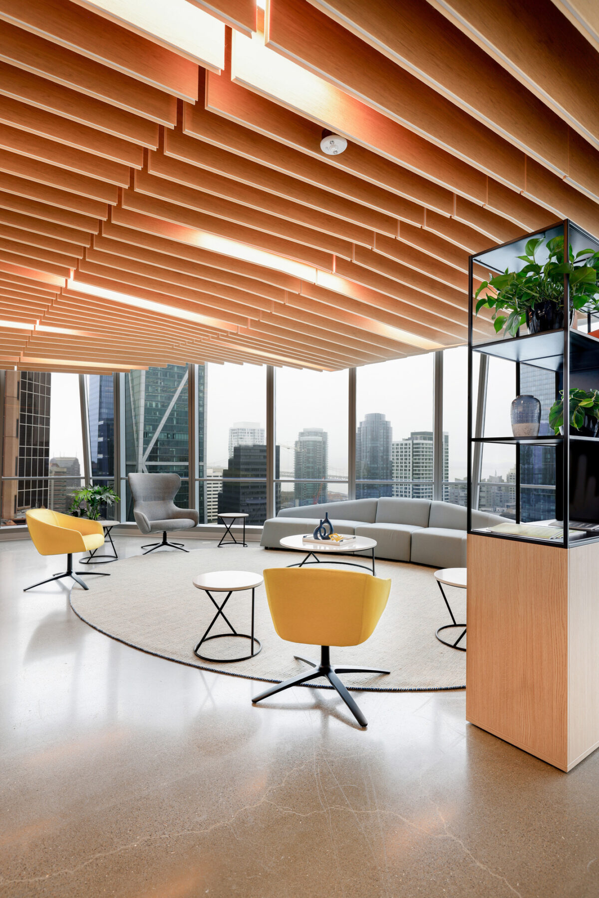 Bright, open workspace featuring a modern aesthetic with a sculptural wooden ceiling. A circular gray rug anchors yellow accent chairs and a minimalist coffee table. Floor-to-ceiling windows provide city views, while potted plants enhance the vibrant atmosphere, blending nature with contemporary design for a collaborative environment.