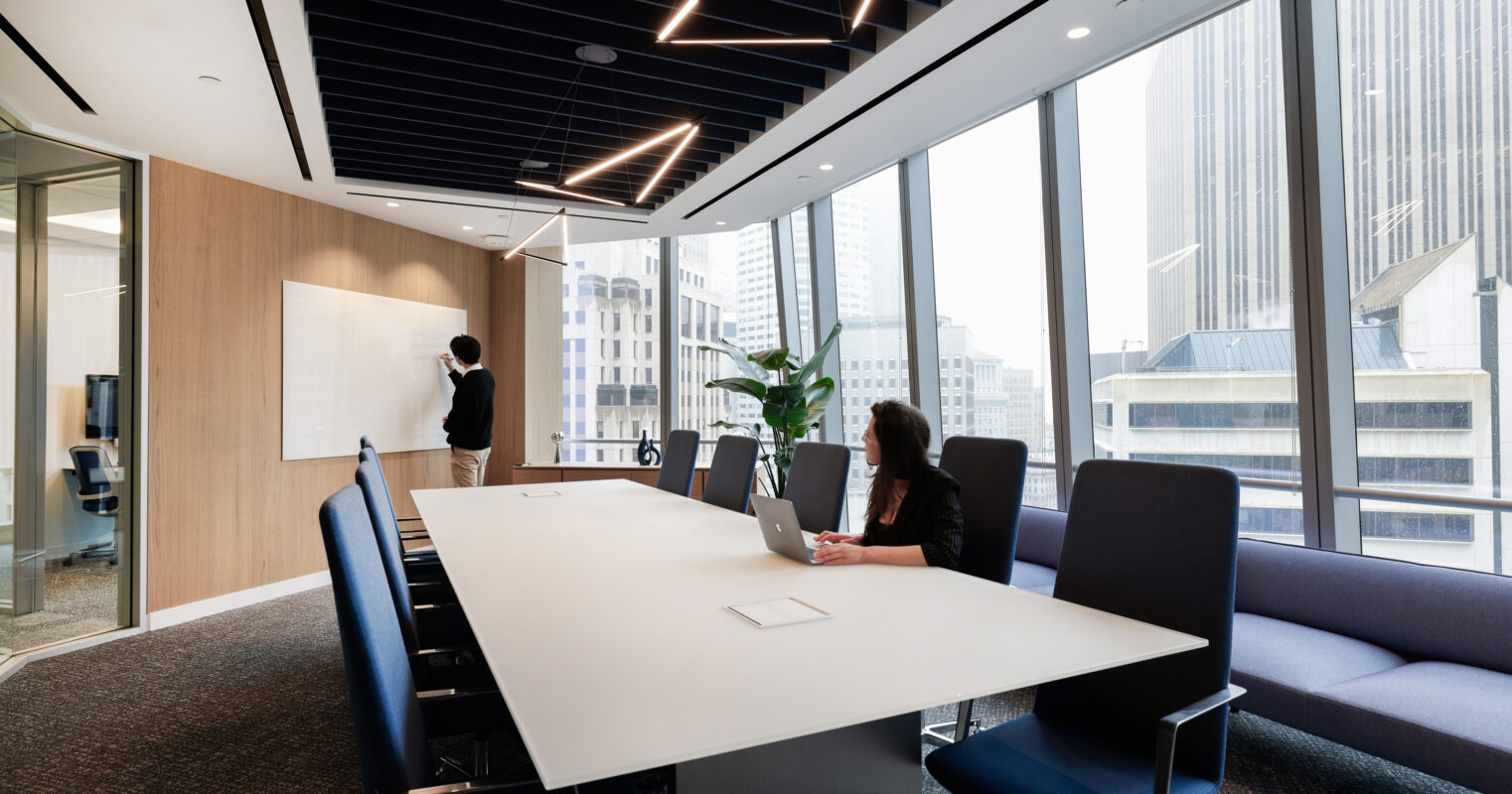 A modern conference room featuring a long, sleek white table surrounded by ergonomic black chairs. A large window showcases a cityscape, providing natural light. Unique architectural details include a dark, slatted ceiling and minimalist light fixtures, creating a contemporary and collaborative atmosphere conducive to meetings.