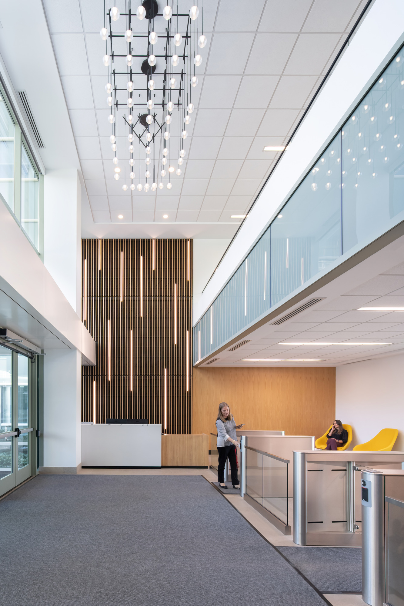 A spacious lobby showcases a modern design with a high ceiling accented by a striking chandelier of clustered lights. Natural wood paneling adds warmth to the contemporary setting, while sleek glass railings enhance openness. Two seating areas feature vibrant yellow chairs, inviting comfort and style in an elegant atmosphere.