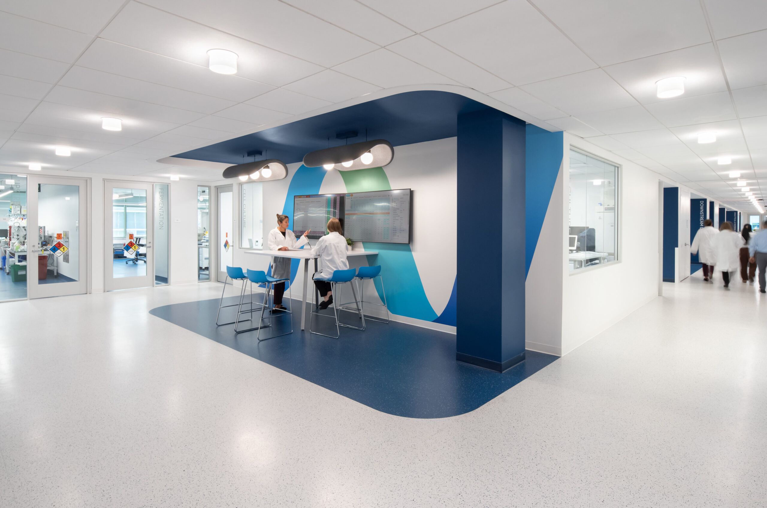 Contemporary healthcare workspace featuring a spacious hallway designed for efficient collaboration. The area showcases a striking blue and green accent wall, complemented by modern circular lighting. Ergonomic seating around a digital display fosters a productive environment for healthcare professionals, enhancing communication and workflow in a clinical setting.
