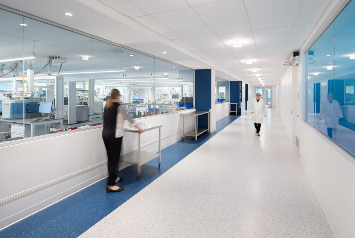 Brightly lit corridor in a modern healthcare facility, featuring expansive glass walls that enhance visibility. The floor showcases a durable, speckled surface in blue and white. Healthcare professionals, dressed in lab attire, navigate seamlessly through the space, promoting an efficient and sterile environment conducive to medical work.