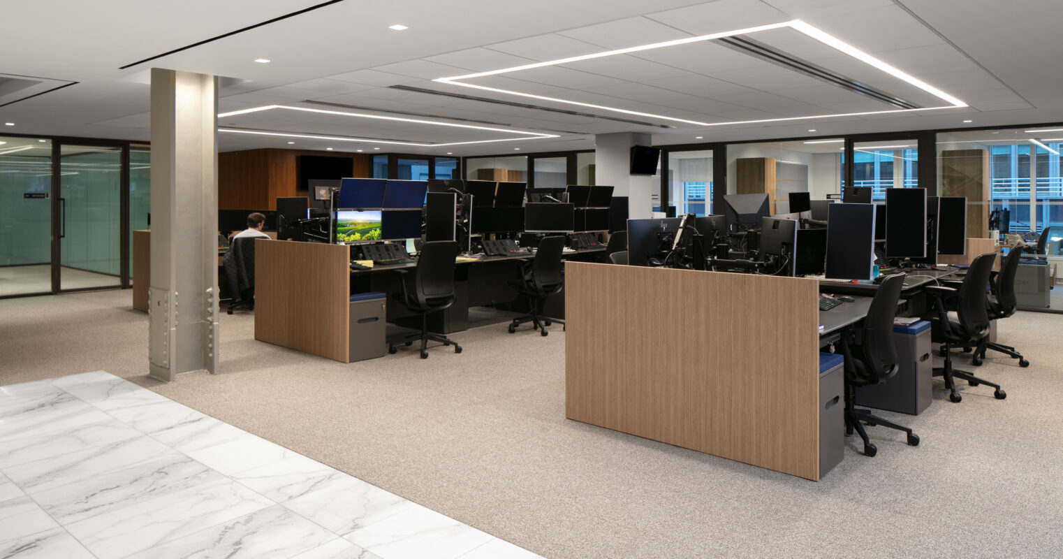 A modern office space featuring a collaborative workstation layout. Warm wooden dividers separate workstations, enhancing privacy while encouraging communication. Large windows provide ample natural light, complemented by soft carpeting and sleek marble tiles, creating a balanced and inviting professional environment that fosters productivity.