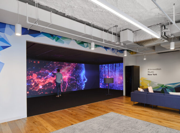 A modern innovation studio featuring dynamic geometric wall graphics in vibrant colors. The space includes a large projection area displaying abstract visuals, fostering creativity and collaboration. Two individuals engage with the technology and ambiance, while contemporary furniture enhances the inviting atmosphere with wooden flooring and textured ceiling elements.