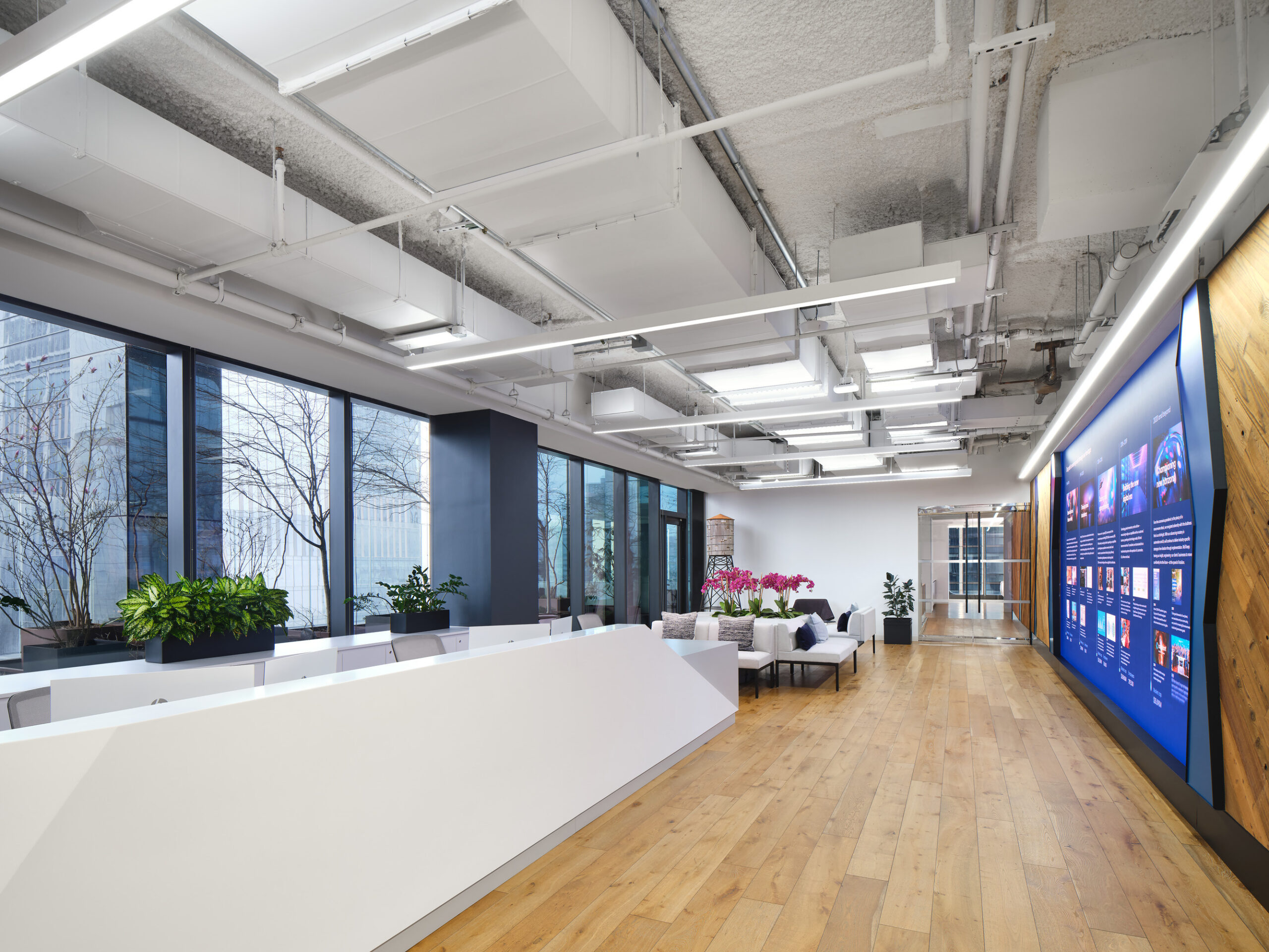 Bright and modern office reception featuring a sleek white front desk and expansive floor-to-ceiling windows that allow natural light to flood the space. The warm wooden flooring contrasts with minimalist furnishings, while lush potted plants and vibrant artwork create a welcoming atmosphere that promotes collaboration and creativity.