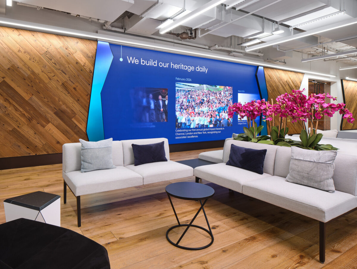 Modern office lounge featuring a blend of natural wood accents and sleek design elements. A vibrant digital display showcases company heritage, while potted orchids add a pop of color. The seating area, with minimalist sofas and a round table, promotes collaboration and comfort in a professional environment.