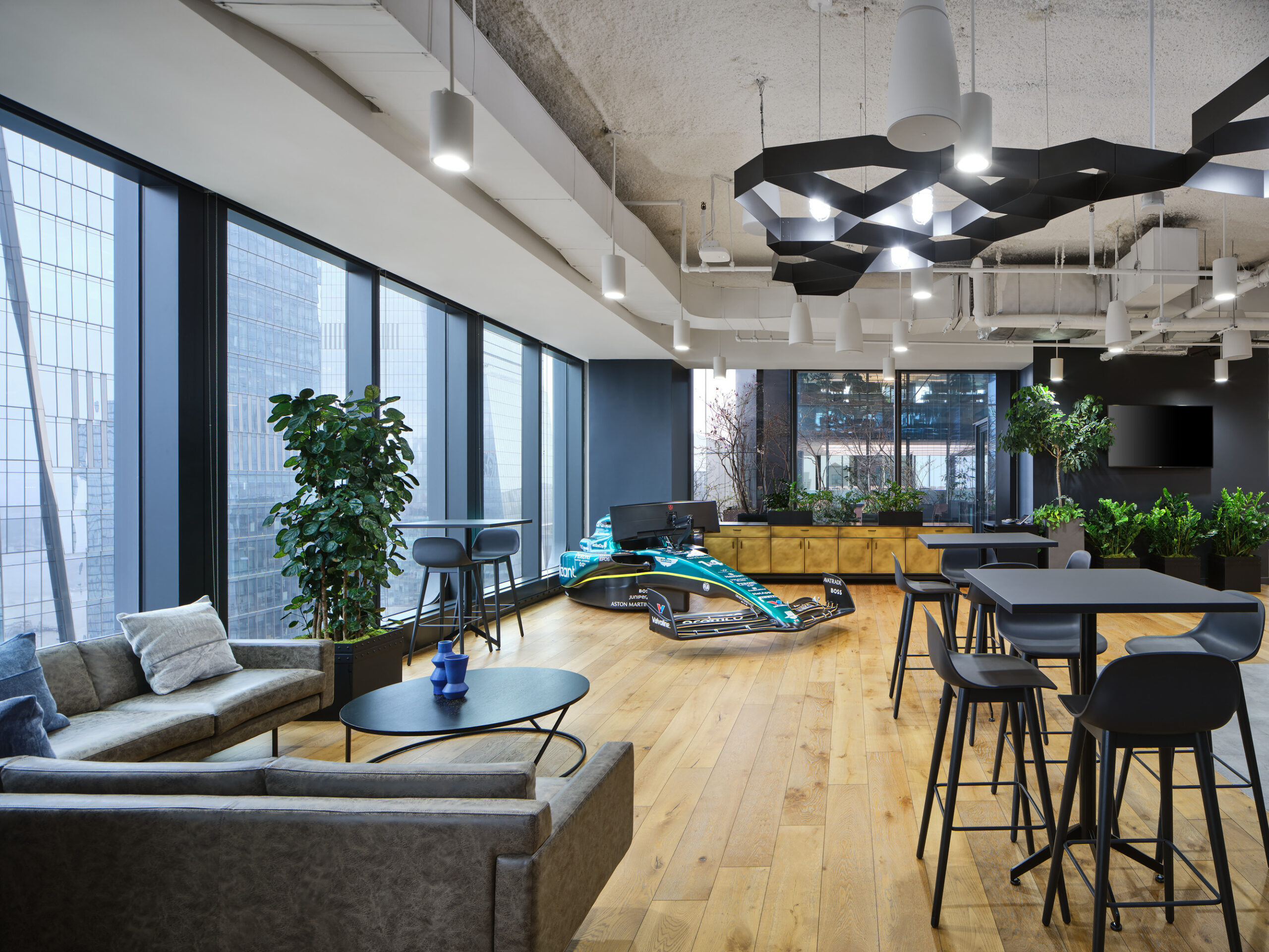 Brightly lit open office space featuring modern furnishings. A plush gray sofa and sleek black tables complement wood flooring. Unique light fixtures hang above while greenery adds life to the area. A futuristic, racing-themed workstation is prominently displayed, enhancing the innovative atmosphere of the workplace.