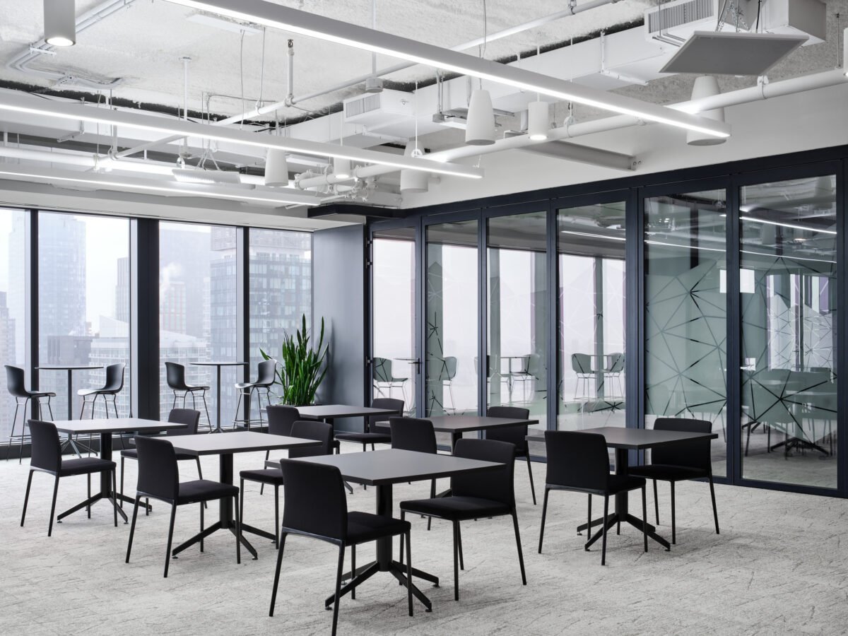 Bright and modern collaborative workspace featuring sleek black modular tables and chairs. Large windows provide ample natural light, offering city skyline views. Glass-walled meeting rooms integrate functionality while geometric wall patterns enhance aesthetic appeal. Minimalist decor emphasizes open space, promoting creativity and interaction among team members.