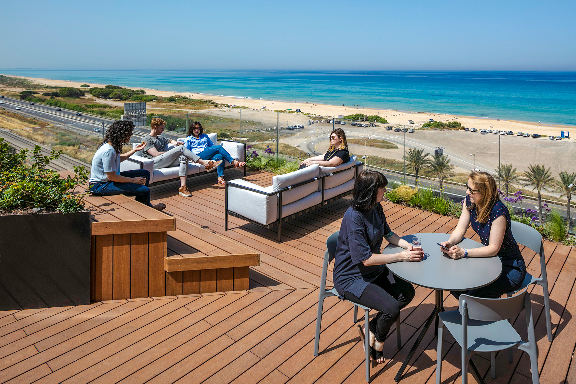 A spacious rooftop terrace featuring modern outdoor furniture, including a sectional sofa and round table. Six people are engaged in conversation, with a panoramic view of the beach and ocean. Lush greenery and wooden decking enhance the inviting atmosphere, creating a relaxing outdoor social space.