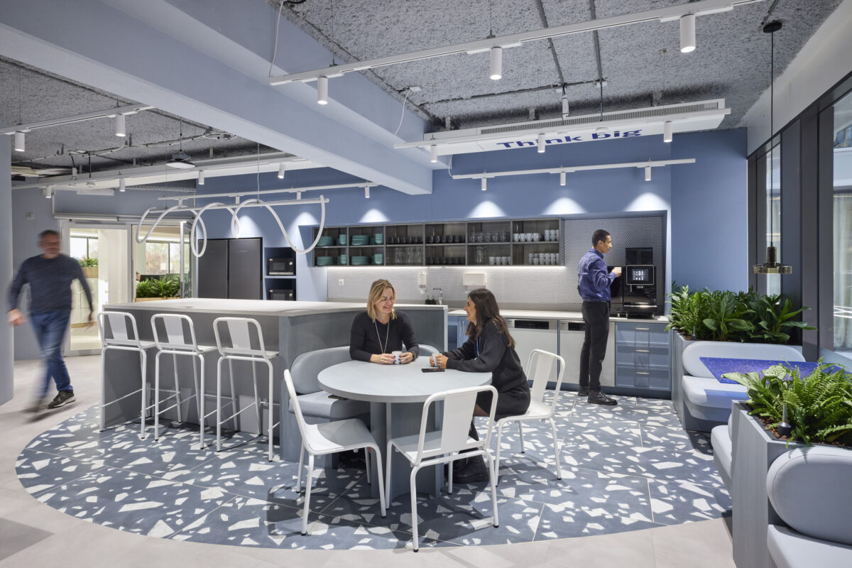 A modern office kitchen featuring a circular table surrounded by white tub chairs on a patterned floor. A sleek, light-gray kitchen island boasts minimalist cabinets and plants. Soft blue walls and elegant lighting create a vibrant, collaborative atmosphere, encouraging conversation and creativity among employees.