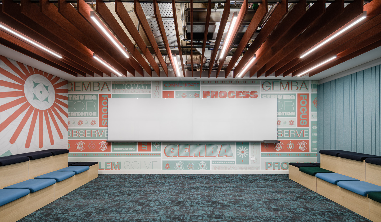 Brightly lit collaborative workspace featuring bold graphic walls with motivational phrases and colored accents. The room is designed with a wooden slatted ceiling and a cozy bench seating area, encouraging teamwork and innovation. The flooring enhances the vibrant atmosphere and fosters a creative environment for meetings and discussions.