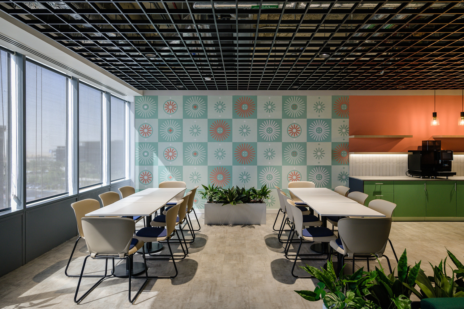 A modern office break room features a contemporary design with a grid-patterned ceiling and large windows allowing natural light. A bold patterned accent wall in pastel colors contrasts with vibrant cabinetry. White tables and chairs are arranged for collaborative use, complemented by greenery in stylish planters, enhancing the inviting atmosphere.