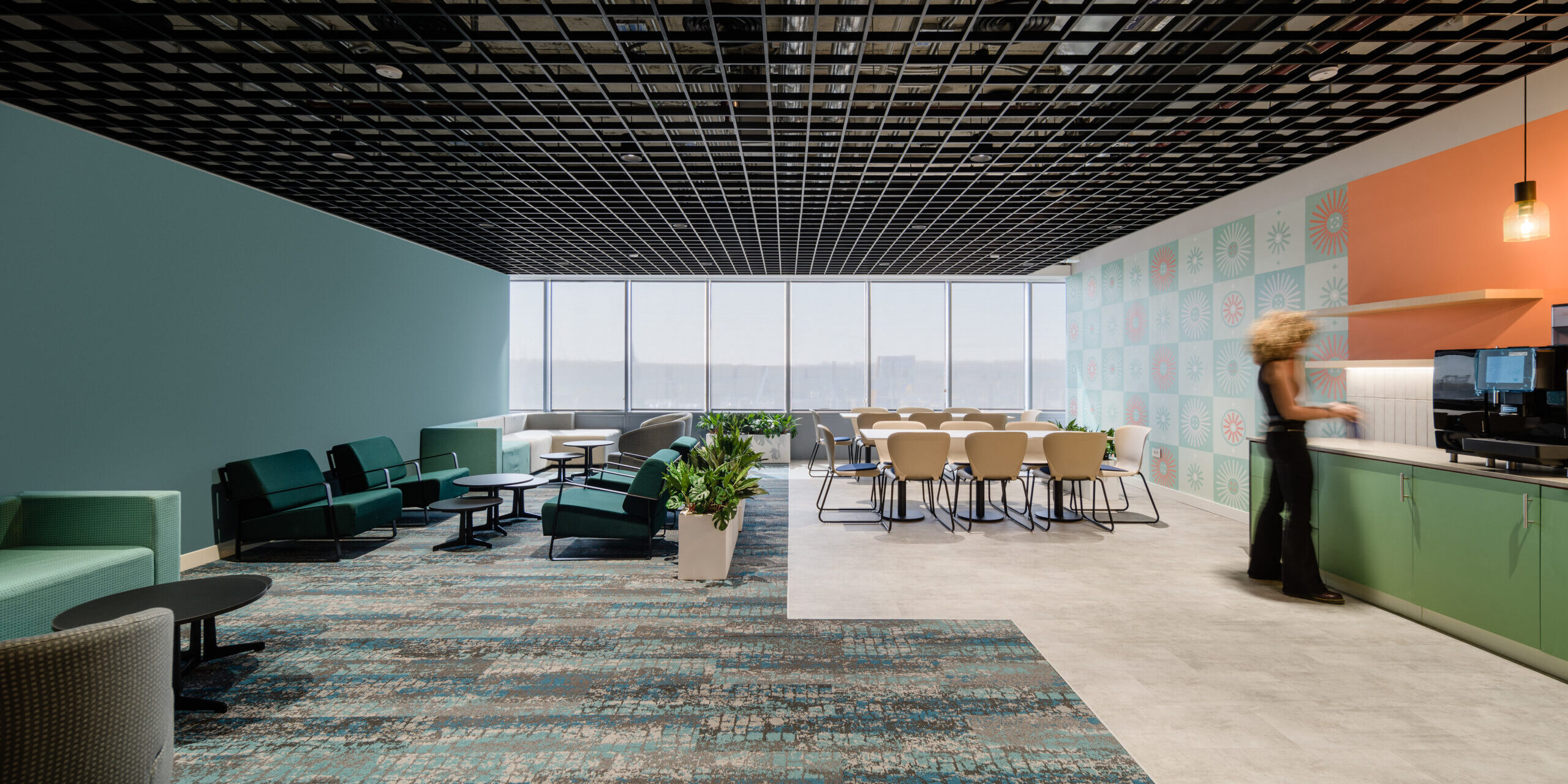 Modern office interior featuring a spacious layout with a grid-patterned ceiling and vibrant wall art. The design incorporates teal and coral accents, complemented by plush seating arrangements and a dining area with light-colored furniture. Lush planters add greenery, enhancing the inviting atmosphere for collaboration and comfort.