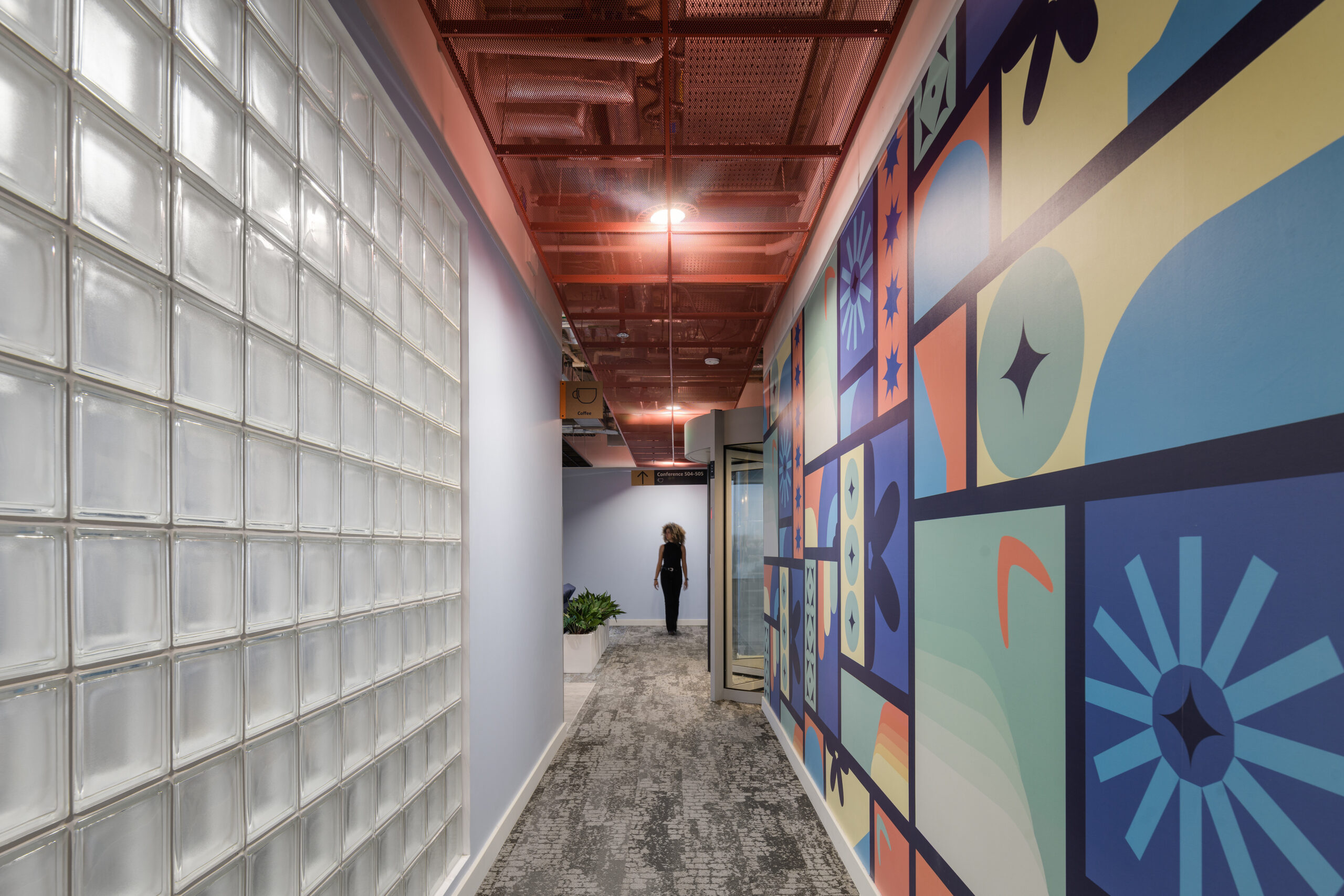 Contemporary corridor featuring a vibrant mural adorned with geometric patterns and bright colors, contrasting against sleek glass block walls. The space is illuminated by modern overhead lighting, complemented by neutral carpeting, creating an inviting yet dynamic atmosphere for office environments. A potted plant adds a touch of nature.