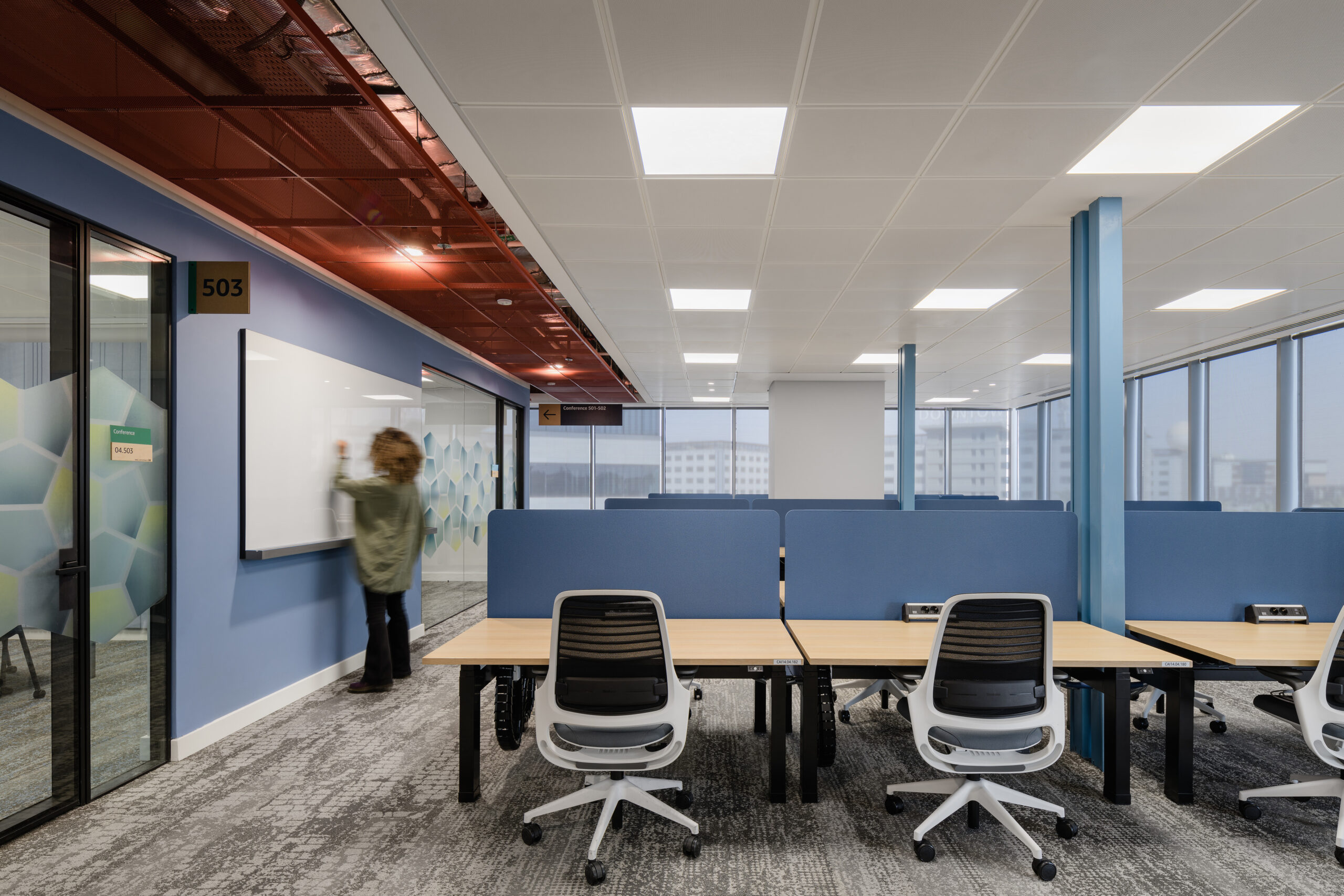 Modern office space featuring collaborative workstations with light wood desks and ergonomic chairs. A person interacts with a whiteboard against a vibrant blue wall, while large windows provide ample natural light. The ceiling showcases a unique red panel design, enhancing the contemporary aesthetic of the workspace.