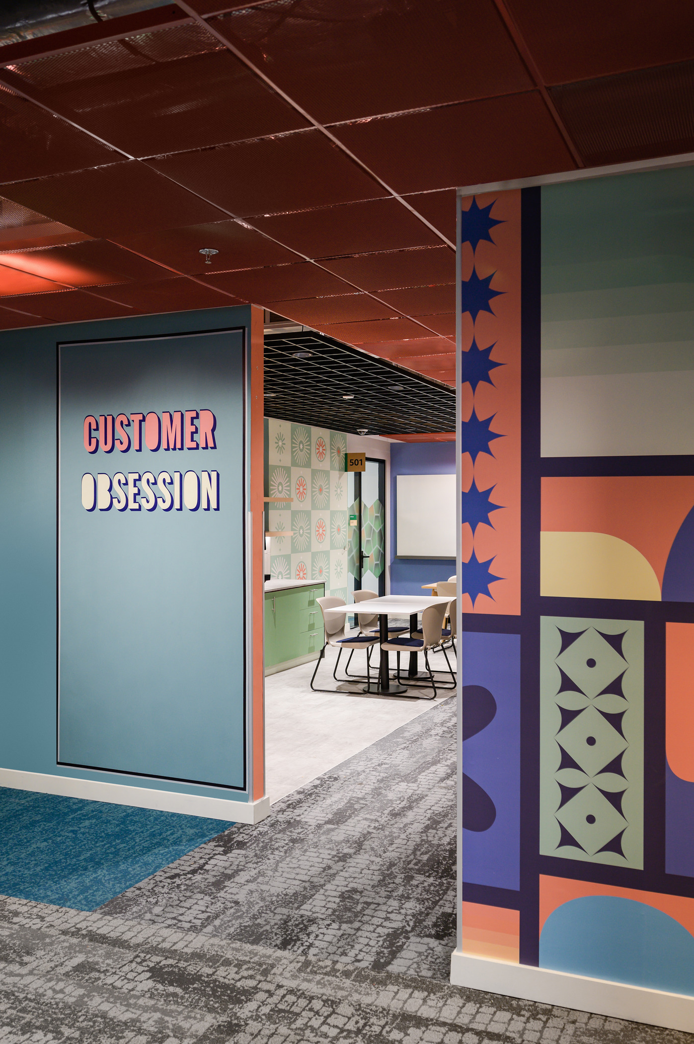 Brightly colored walls feature intricate geometric patterns and playful typography, creating an engaging atmosphere in a modern office space. The "CUSTOMER OBSESSION" sign adds a focal point, while the adjacent meeting area showcases a minimalist table and chairs against a vibrant backdrop, emphasizing creativity and collaboration.