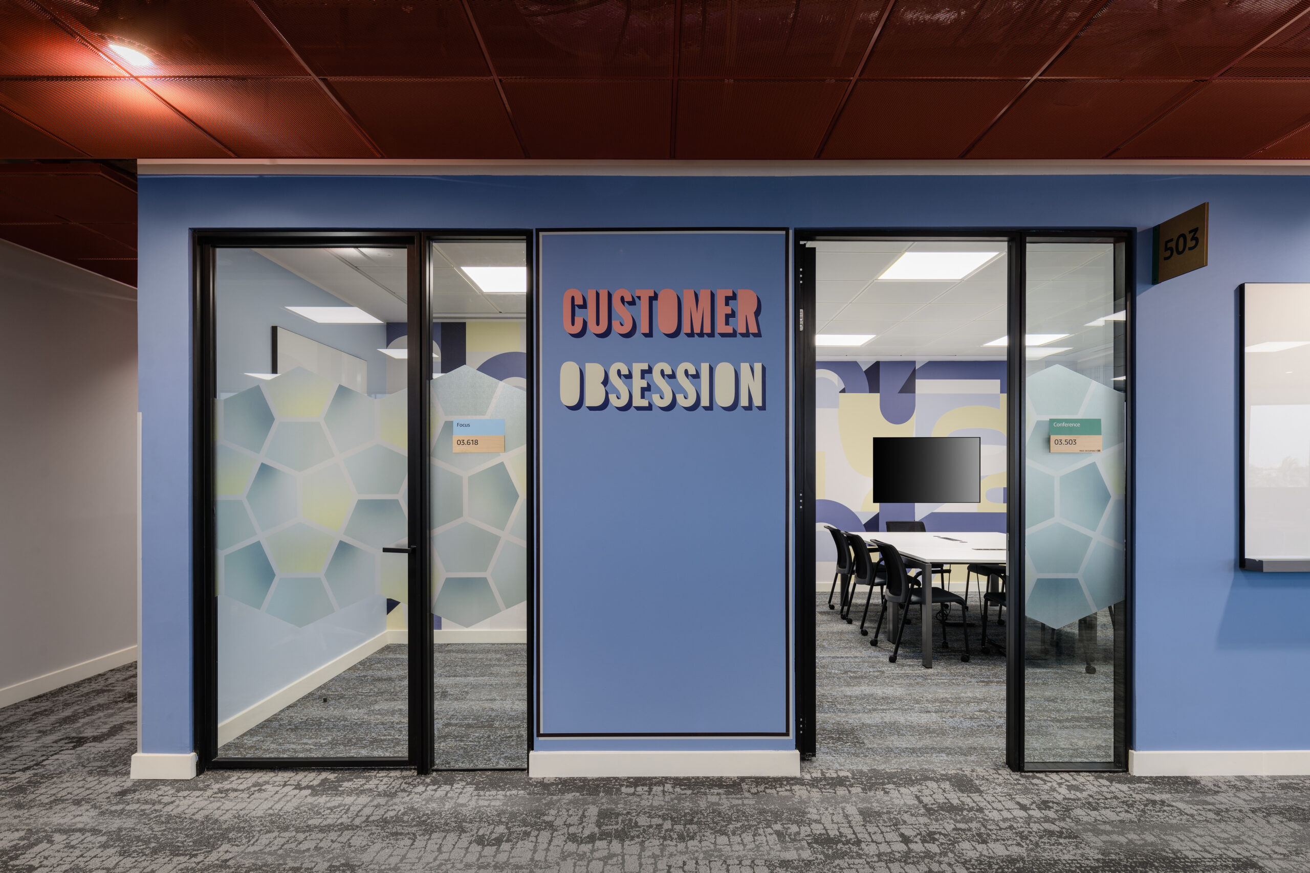 Brightly colored office space features a glass-walled meeting room labeled "Customer Obsession." The engaging blue and patterned walls create a vibrant atmosphere, while hexagonal graphics on the glass panels enhance the modern design. The neutral carpet and ceiling add balance, promoting a stylish yet functional workspace.