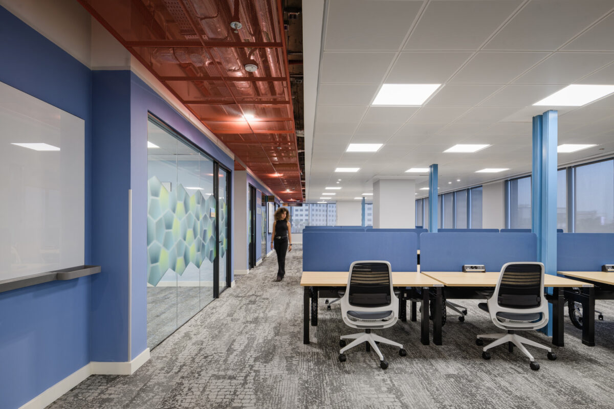 Brightly lit modern office corridor featuring sleek, open workspaces with ergonomic chairs. The walls are painted in a soft blue, complemented by a vibrant red ceiling. Glass-walled conference rooms showcase hexagonal patterns, enhancing collaboration. Large windows allow natural light to flood the space, creating an inviting and productive environment.