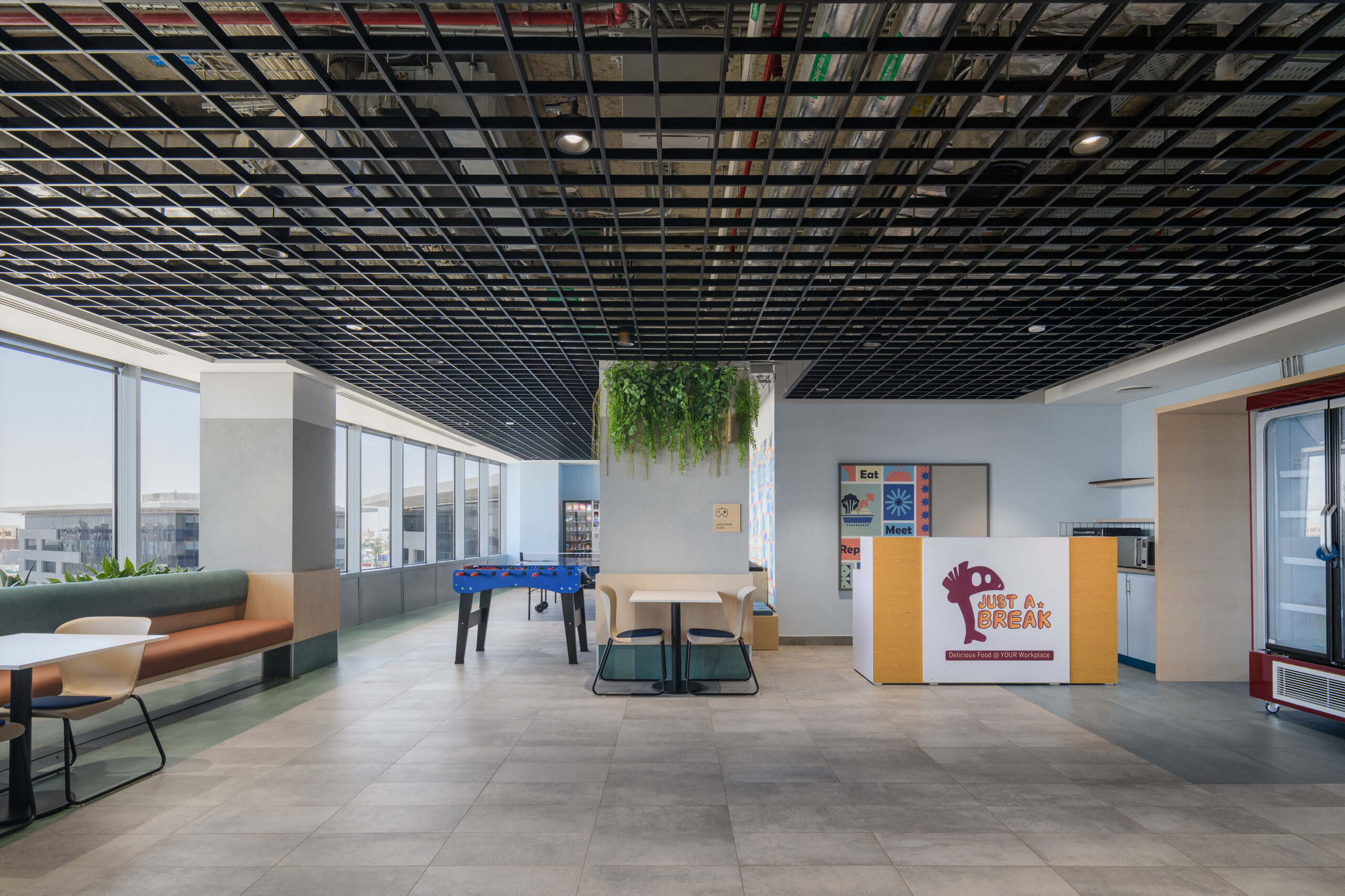 A modern office break area features a striking black grid ceiling reflected in large windows, allowing ample natural light. Soft seating in neutral tones complements playful design elements like a foosball table and a vibrant mural. Greenery adds a refreshing touch to the contemporary interior.