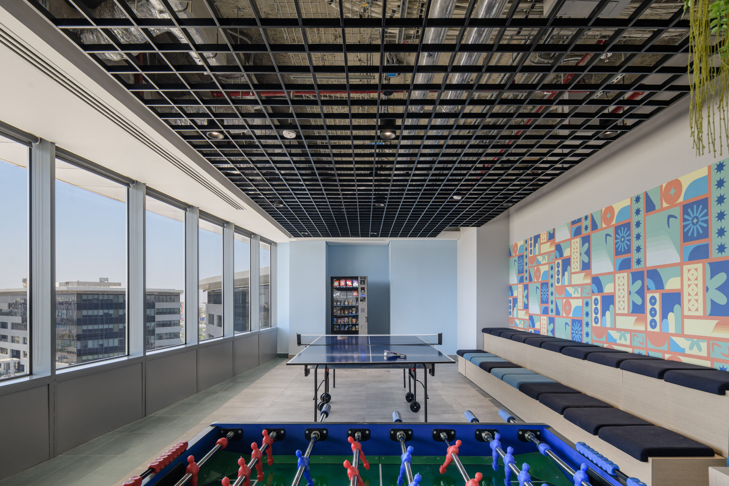A modern office break area featuring expansive windows that provide natural light. A sleek black grid ceiling contrasts with a vibrant, patterned mural on one wall. A multifunctional space accommodates a foosball table and ping pong table, with minimalistic seating along the side, promoting relaxation and collaboration.