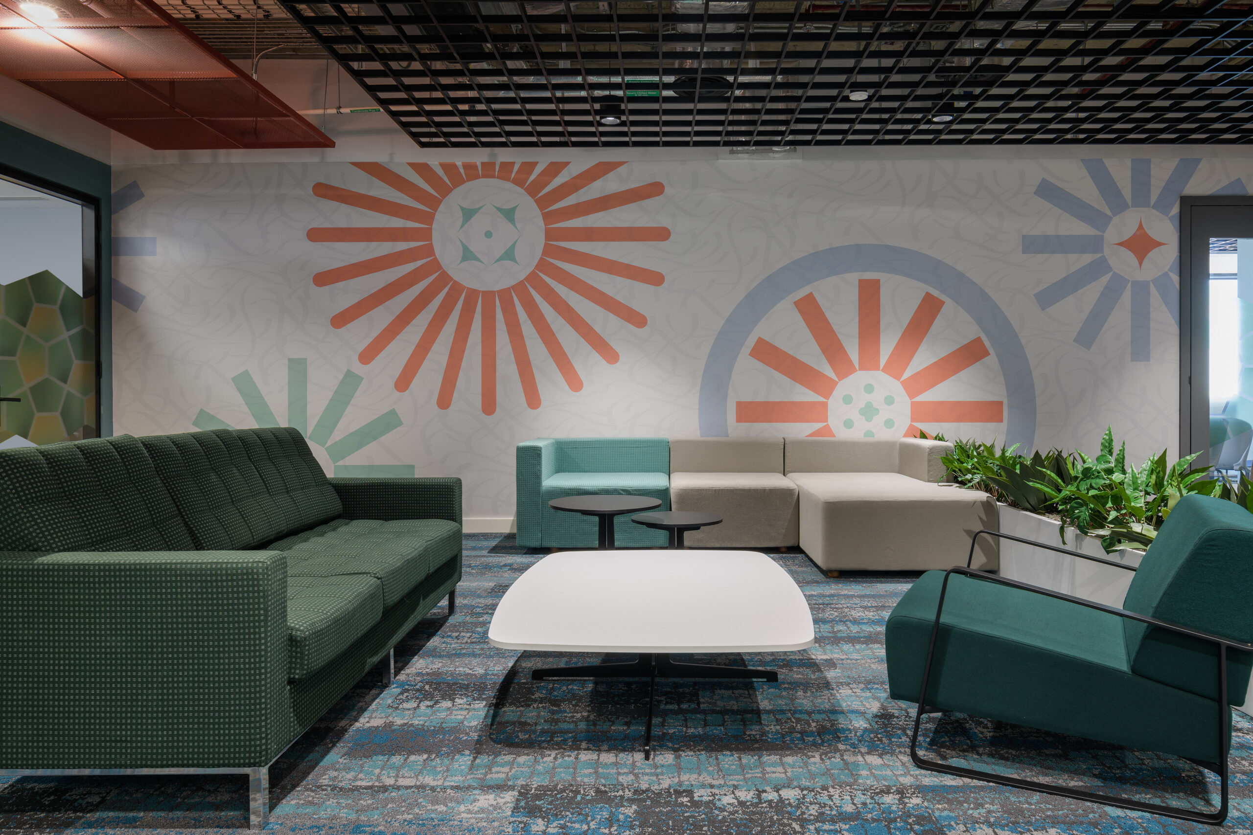 A contemporary lounge area features a mix of textured seating, including a green sectional, modular sofa, and vibrant accent chairs. A white coffee table centers the space, surrounded by artistic wall murals with geometric patterns and colors that enhance the inviting atmosphere, complemented by strategically placed greenery.
