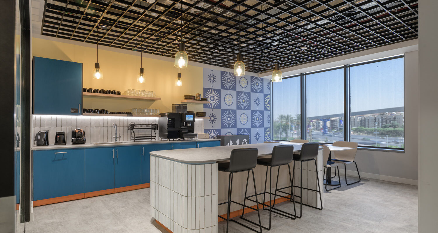 This modern kitchenette features a striking black grid ceiling that contrasts with the bright blue cabinetry. The well-organized space includes a coffee machine and an elegant breakfast bar with six stylish high stools. Large windows allow natural light, accentuating the playful wall design adorned with geometric patterns.