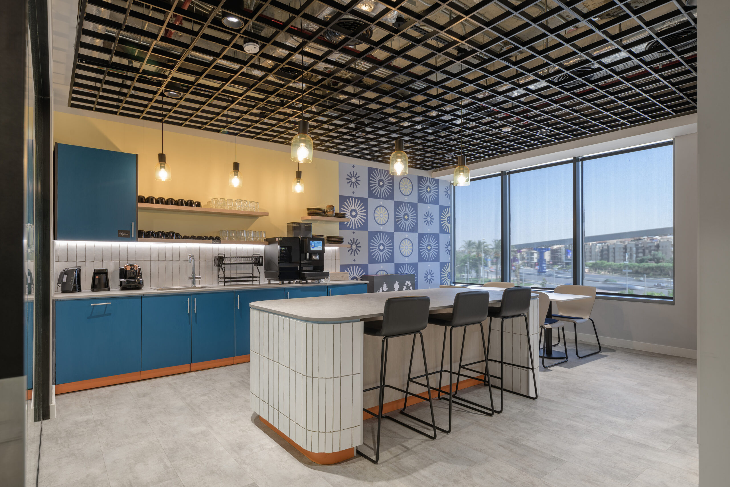 Modern break room featuring a contemporary kitchen design with bold blue cabinets and a striking patterned tile backsplash. An island with a subtle curved edge provides seating with sleek black bar stools. Illuminated by pendant lights, large windows offer ample natural light and city views.