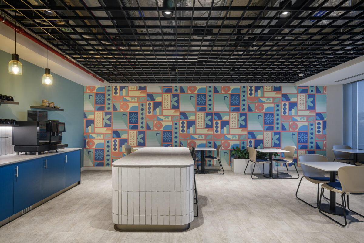 A modern office break room featuring vibrant, abstract wallpaper in pastel colors. The space includes a sleek, white island with a textured countertop, blue cabinetry, and contemporary seating. The grid-patterned ceiling enhances the stylish ambiance, while pendant lighting adds warmth and sophistication to the design.