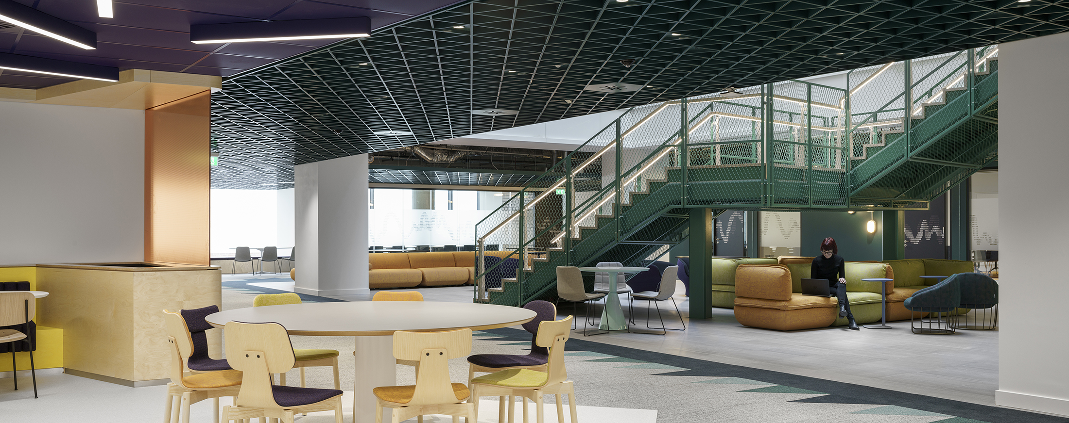 A contemporary open-plan interior showcasing a vibrant lounge area with green and gold accents. The design features a modern staircase leading to elevated seating areas, complemented by colorful chairs and a mixed-use table. Sleek geometric ceilings enhance the space, promoting a collaborative and inviting atmosphere.