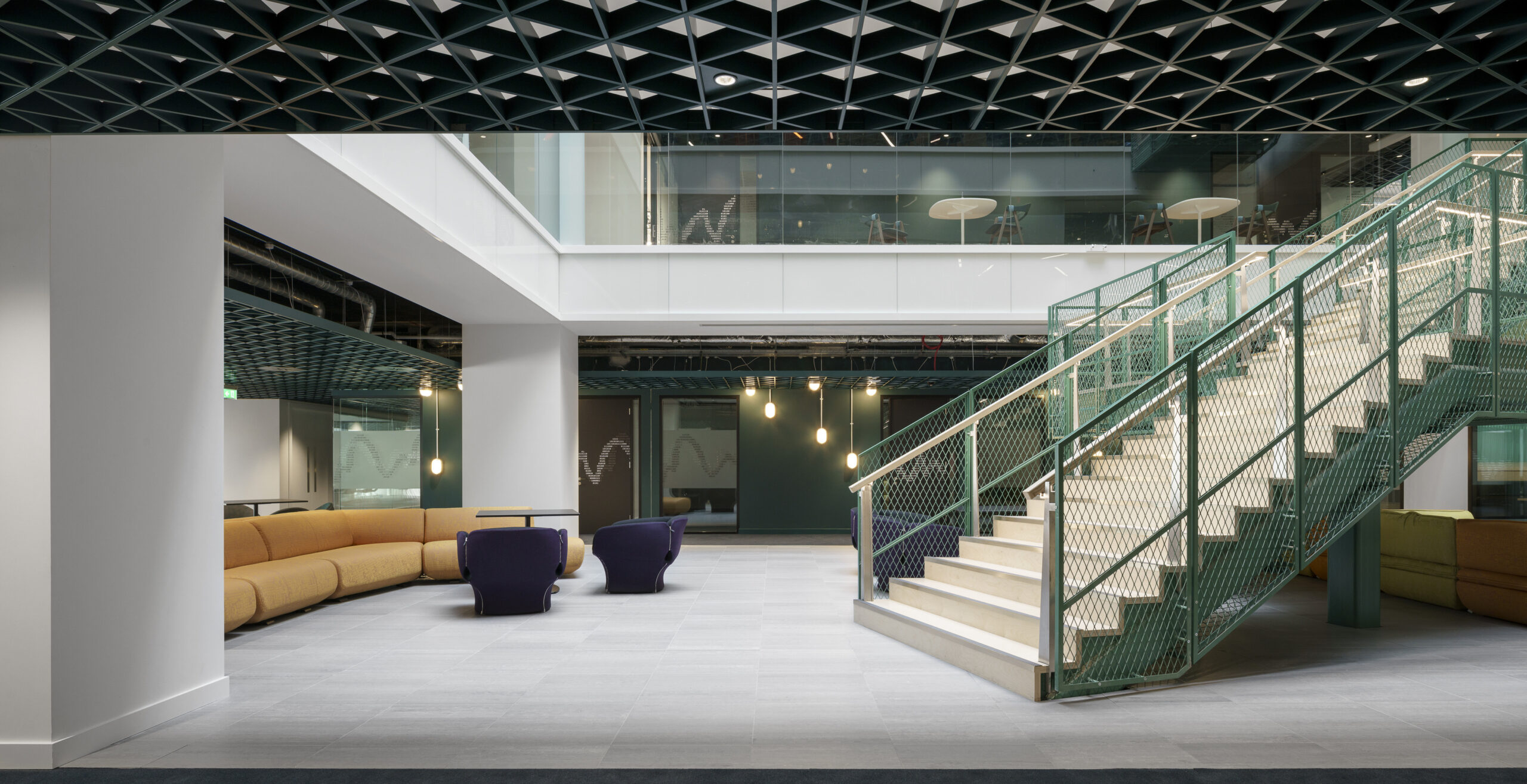 A spacious, contemporary lobby features a strikingly geometric ceiling and a mix of textures. Curved seating in warm tones contrasts with sleek, modern furniture in dark blue. A prominent staircase with a teal metal railing invites exploration, illuminated by soft, adjustable pendant lighting, enhancing the open, inviting atmosphere.