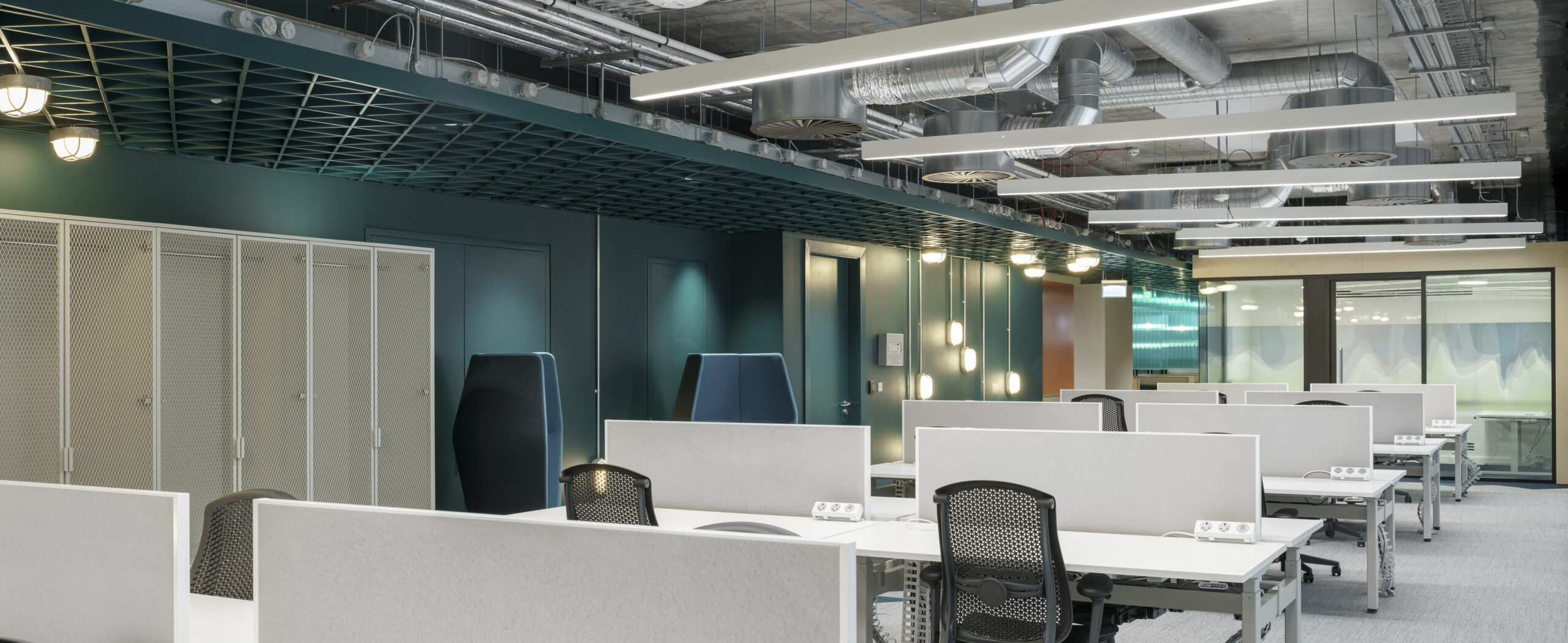 Modern office interior featuring open workspaces designed for collaboration. Light gray workstations equipped with ergonomic chairs are arranged in a grid layout, accented by sleek black ceilings and industrial piping. Soft lighting from overhead fixtures enhances the professional atmosphere, promoting productivity and comfort. Private phone booths provide quiet spaces for focused work.