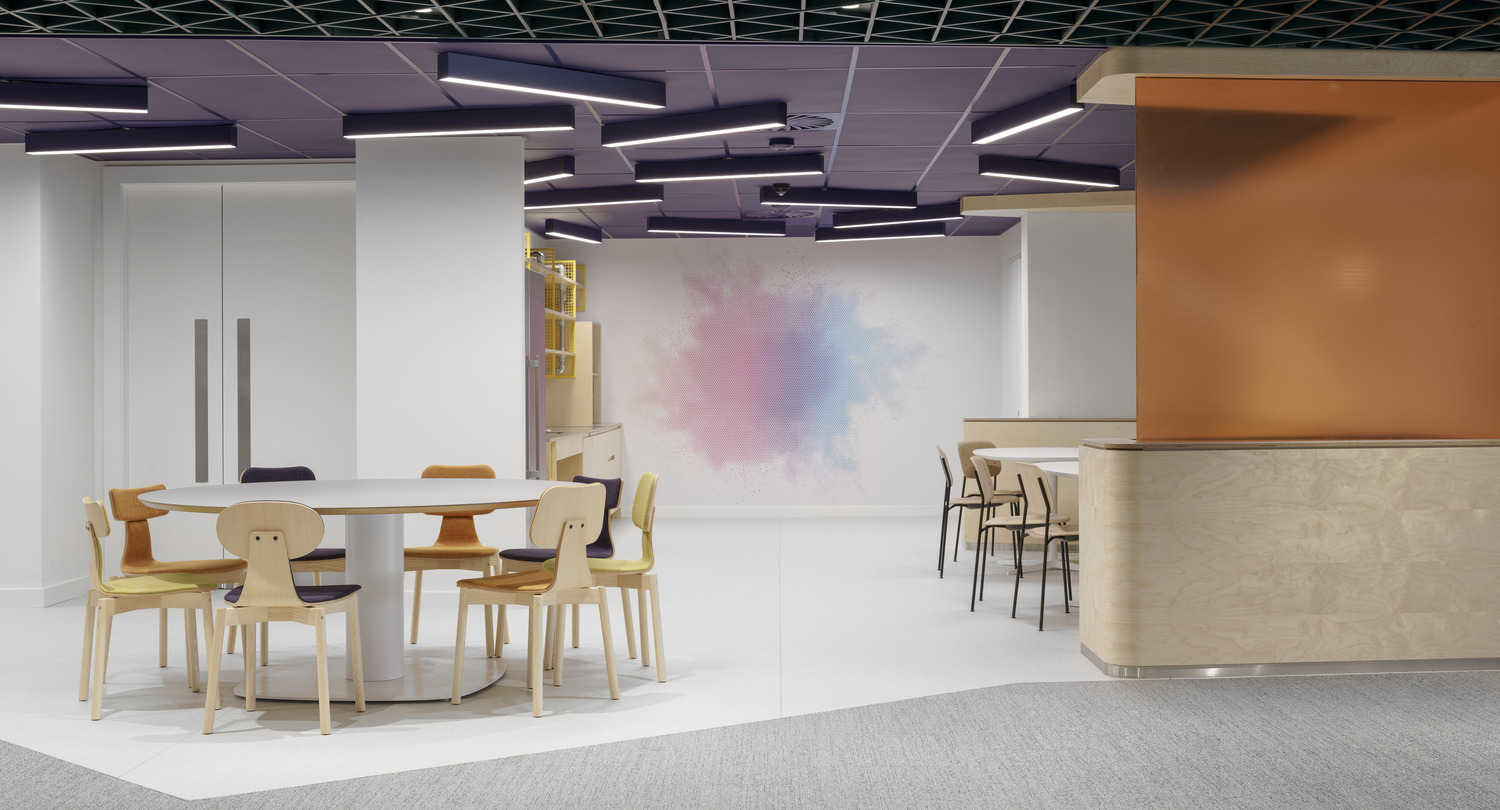 Brightly lit collaborative workspace featuring a round white table surrounded by colorful, mismatched chairs. A textured wall with a soft, pastel splash serves as a focal point, while modern pendant lighting with geometric shapes creates a dynamic ceiling feature. The space combines functionality with playful design elements.