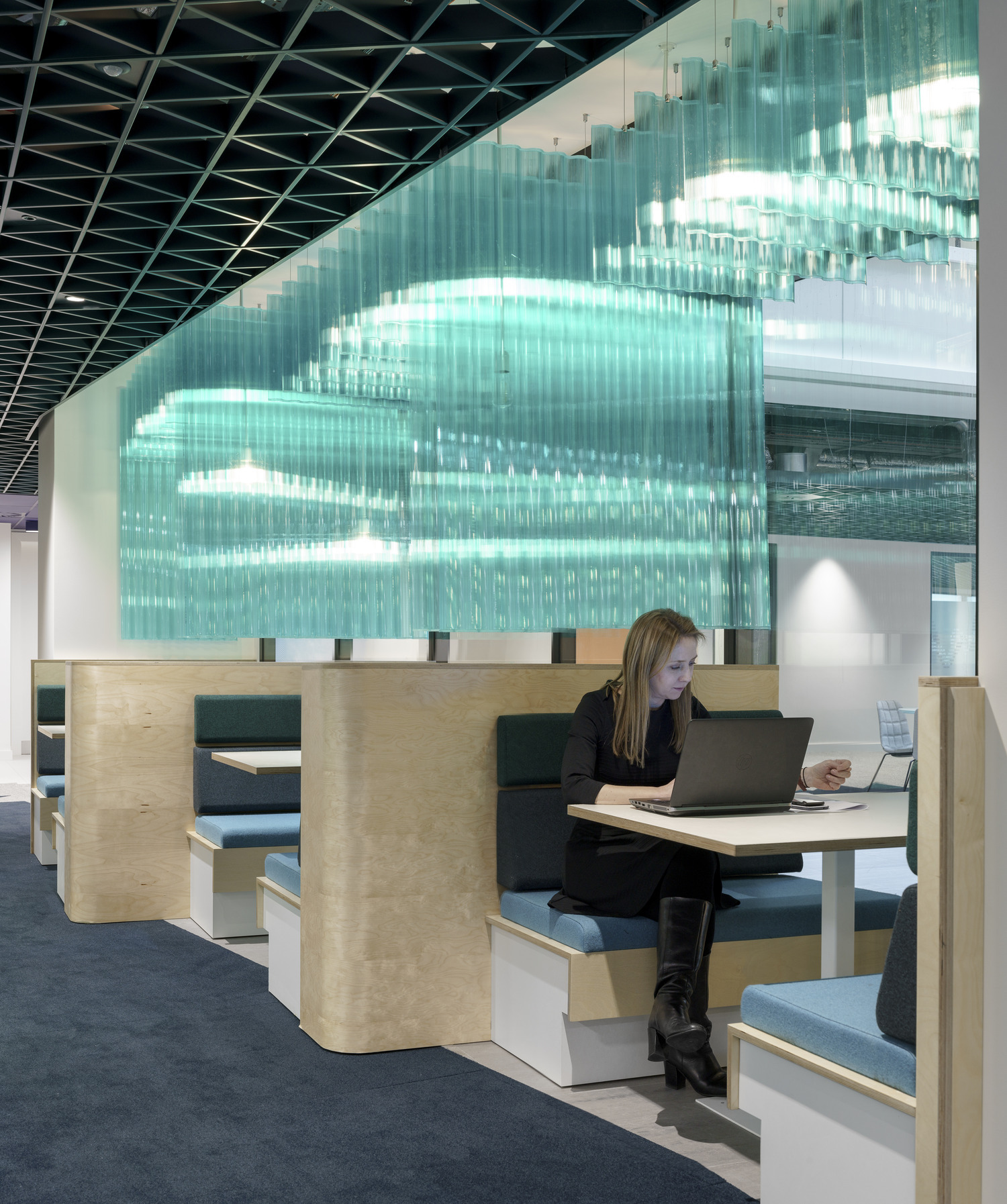 Contemporary workspace featuring modular booths with light wood finishes and upholstered seating in soft hues. A professionally dressed woman is engaged in work at a sleek table, illuminated by an eye-catching, translucent glass wall that creates a dynamic light effect, enhancing the overall creative atmosphere of the office.