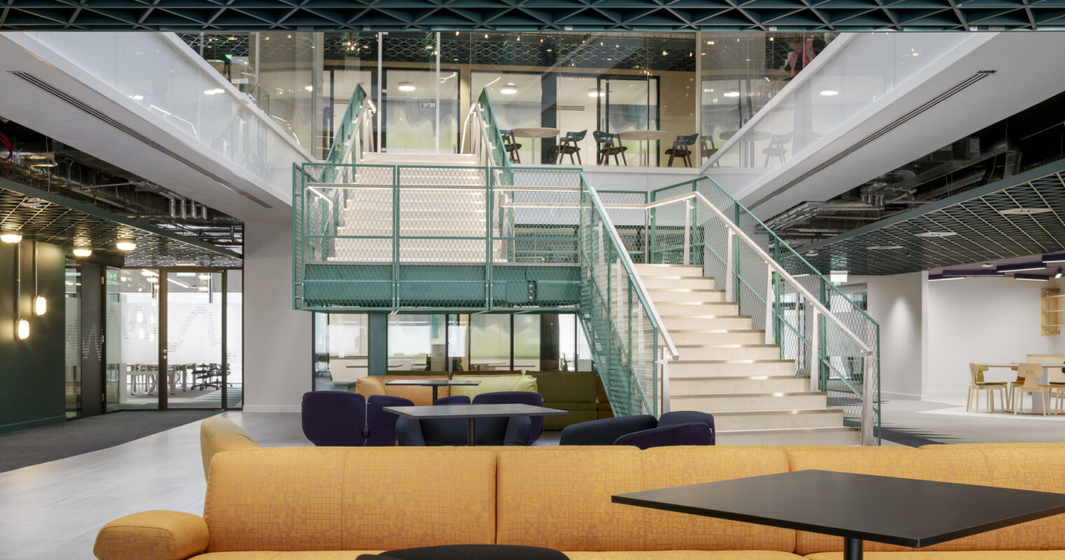 A spacious, modern interior features an open atrium with a glass staircase and multiple seating areas. Brightly colored furniture, including a yellow sectional sofa and dark lounge chairs, complements the sleek architectural elements and large windows, promoting a collaborative and inviting environment for occupants.