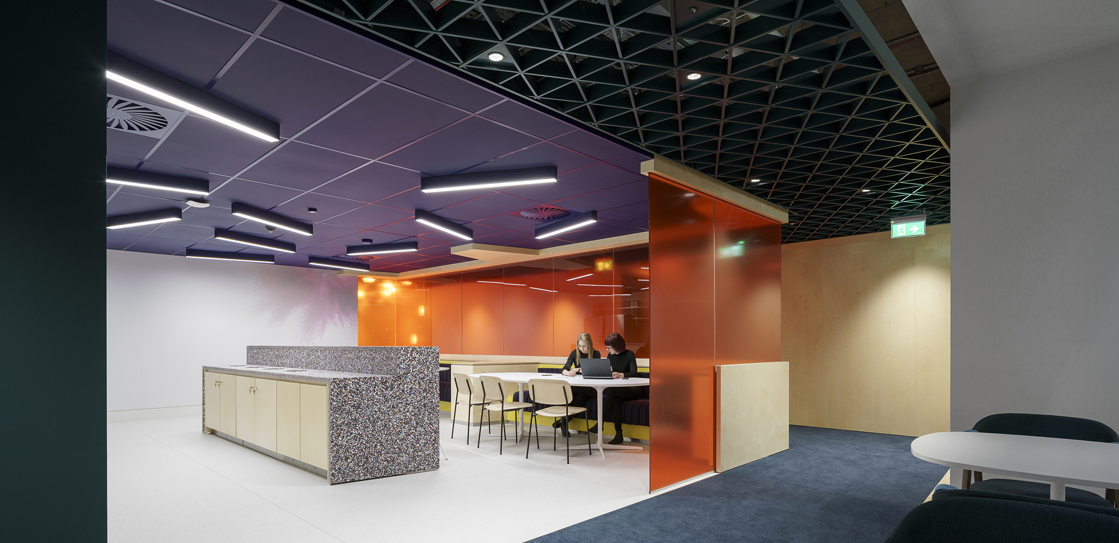 A modern office collaboration space featuring a vibrant orange partition that contrasts with the minimalist design. The ceiling is adorned with a geometric grid of black tiles, while fluorescent light fixtures provide illumination. A communal table with multi-colored chairs encourages teamwork and creativity within the workspace.