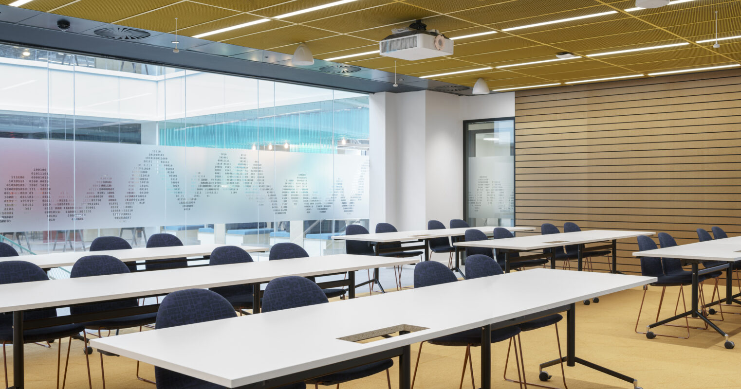 Modern classroom featuring flexible seating arrangements with white tables and blue ergonomic chairs. Transparent glass walls promote natural light while offering a view into the collaborative space. Acoustic panels on the right enhance sound quality, and integrated technology including a projector indicates a focus on innovative learning practices.