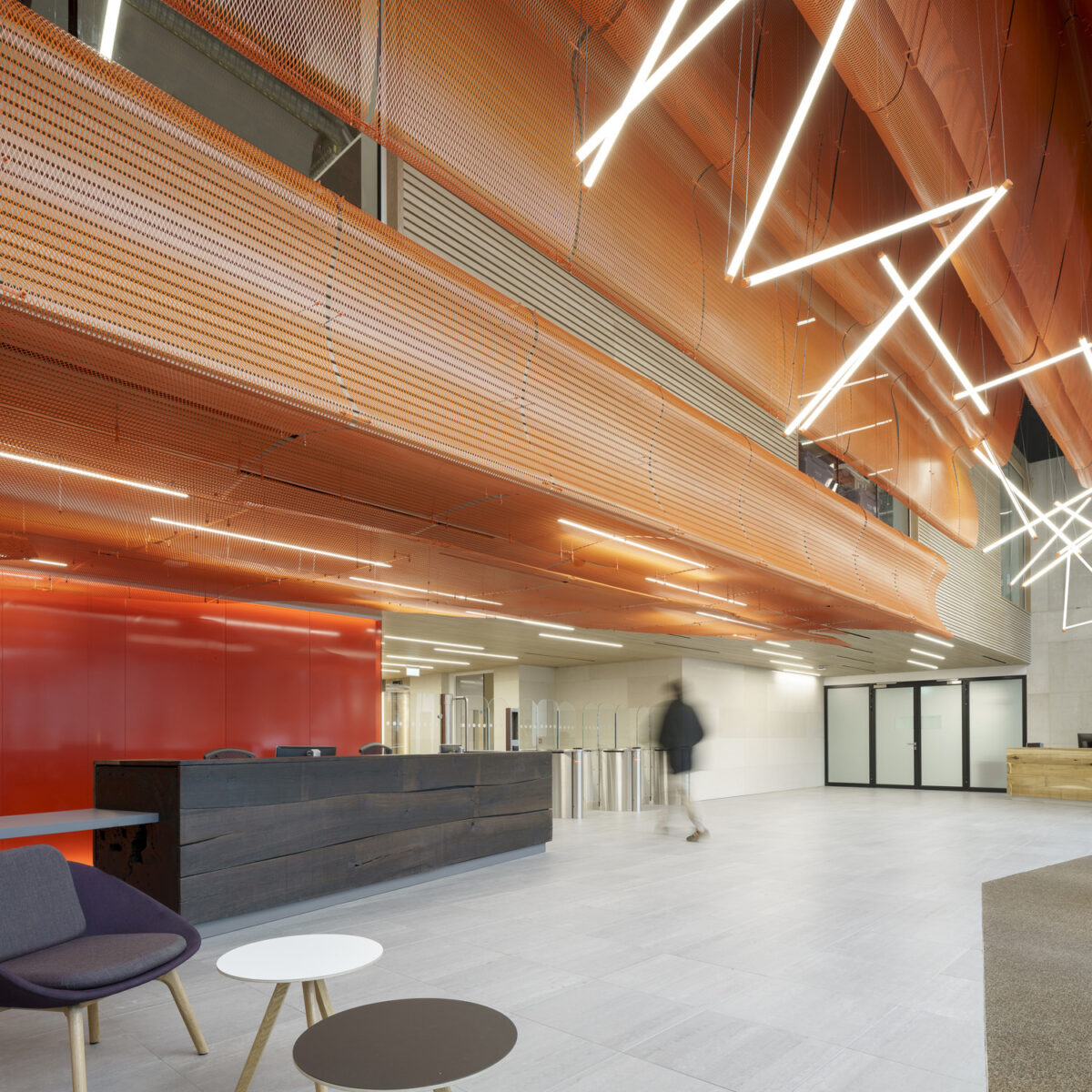 Brightly lit reception area featuring a striking ceiling design with interwoven orange panels and angular lighting fixtures. Minimalist furniture includes a gray armchair and small round tables in contrasting colors. The sleek, modern aesthetic is complemented by a polished stone floor and a vibrant red accent wall.