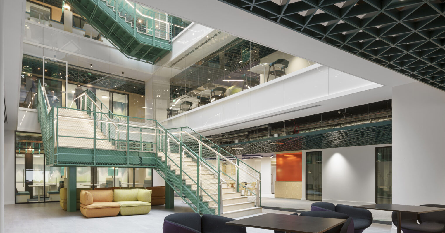 A spacious, modern atrium featuring a striking green geometric ceiling. The area is illuminated by abundant natural light filtering through large windows. A prominent staircase with a unique railing leads to upper levels, while colorful, contemporary seating areas invite collaboration and relaxation, showcasing a blend of functionality and aesthetics in interior design.