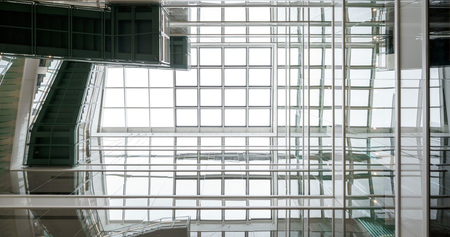 A modern atrium features a striking glass ceiling, allowing natural light to permeate the space. The interplay of transparent and reflective surfaces creates a dynamic visual experience, emphasizing the architectural lines while enhancing the spaciousness of the interior. Green accents add a touch of nature to the design.