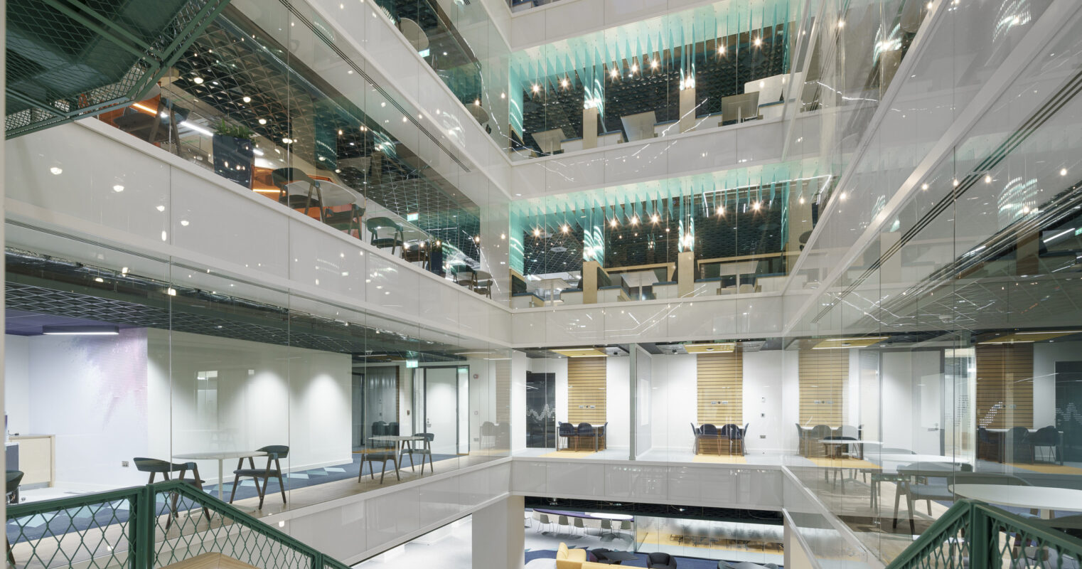 Open atrium design featuring modern glass and metal elements, showcasing interconnected office spaces. The multi-level layout is illuminated by ambient lighting, enhancing the contemporary feel. Striking green accents in mesh overhead structures complement the elegant wooden staircase, creating a dynamic, collaborative workspace atmosphere.