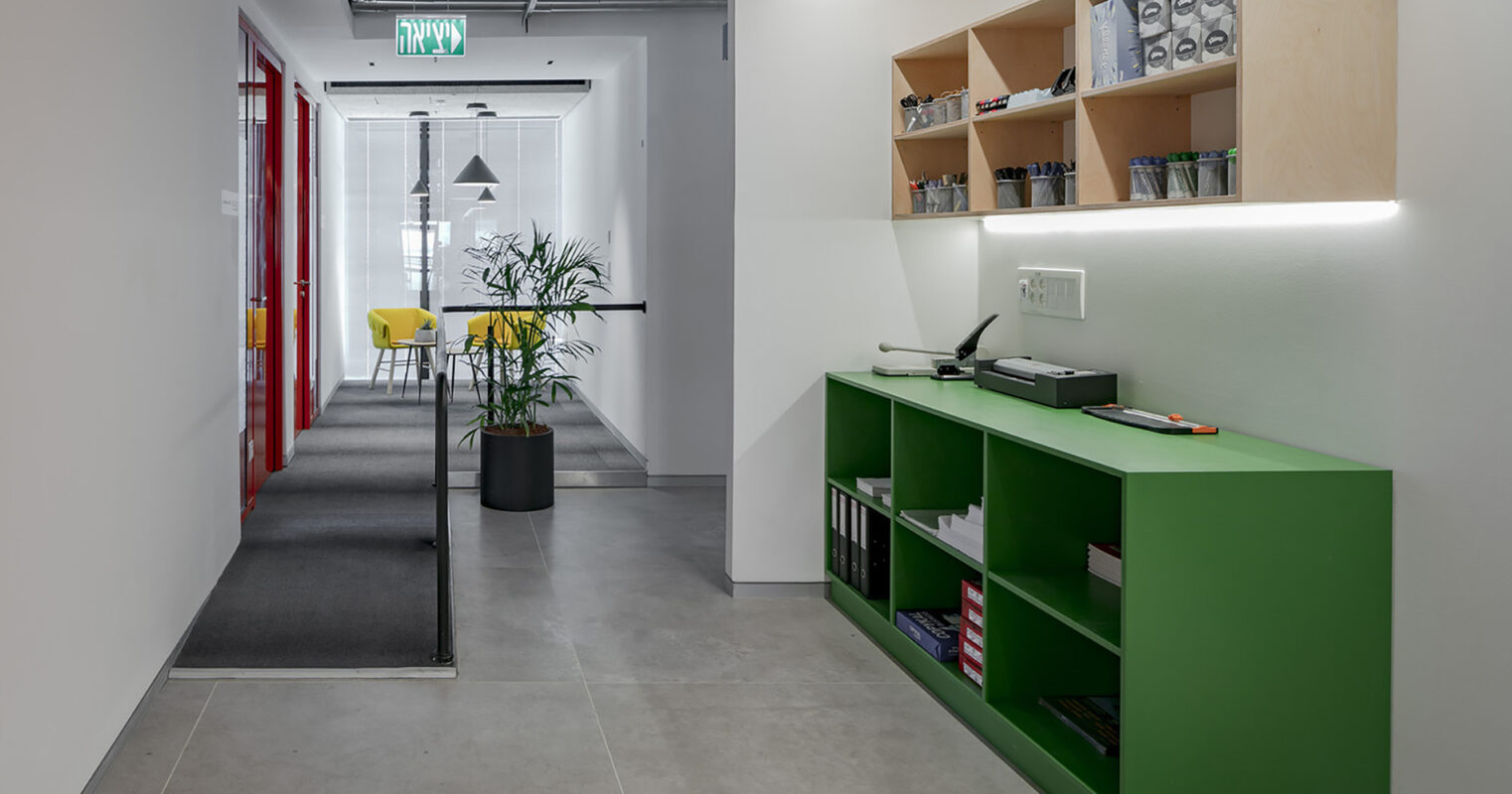 Brightly lit entrance hall showcasing a minimalist design. A sleek green console table with open shelving contrasts against the neutral walls. To one side, a plant adds a touch of greenery, while in the distance, a meeting area features vibrant yellow chairs, creating an inviting atmosphere.