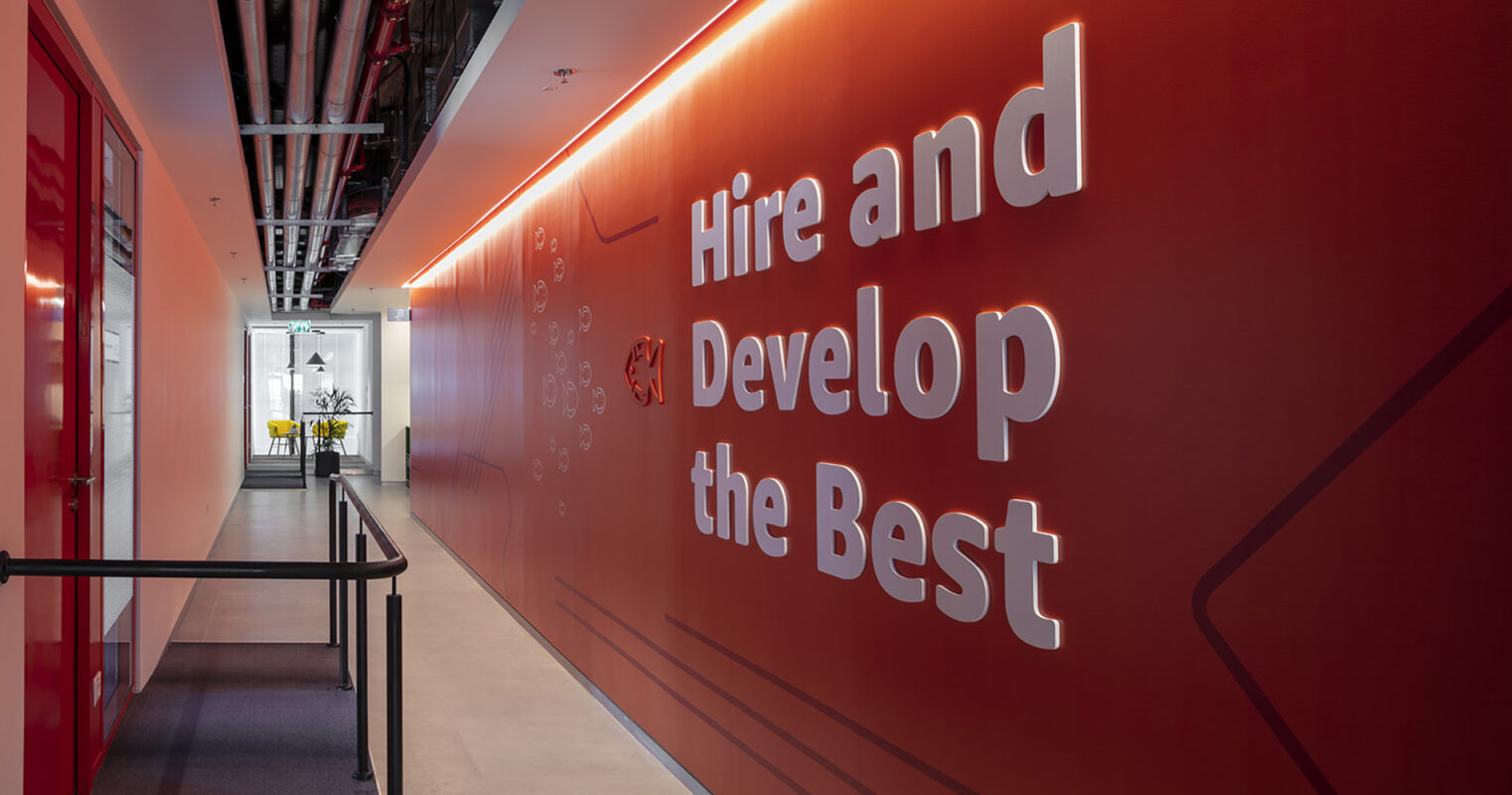 Bold, illuminated text on a vibrant orange wall reads “Hire and Develop the Best.” The modern corridor features smooth flooring and sleek railings, enhancing the dynamic atmosphere. Soft, ambient lighting complements the design, creating an inviting workspace that emphasizes innovation and motivation in an office setting.