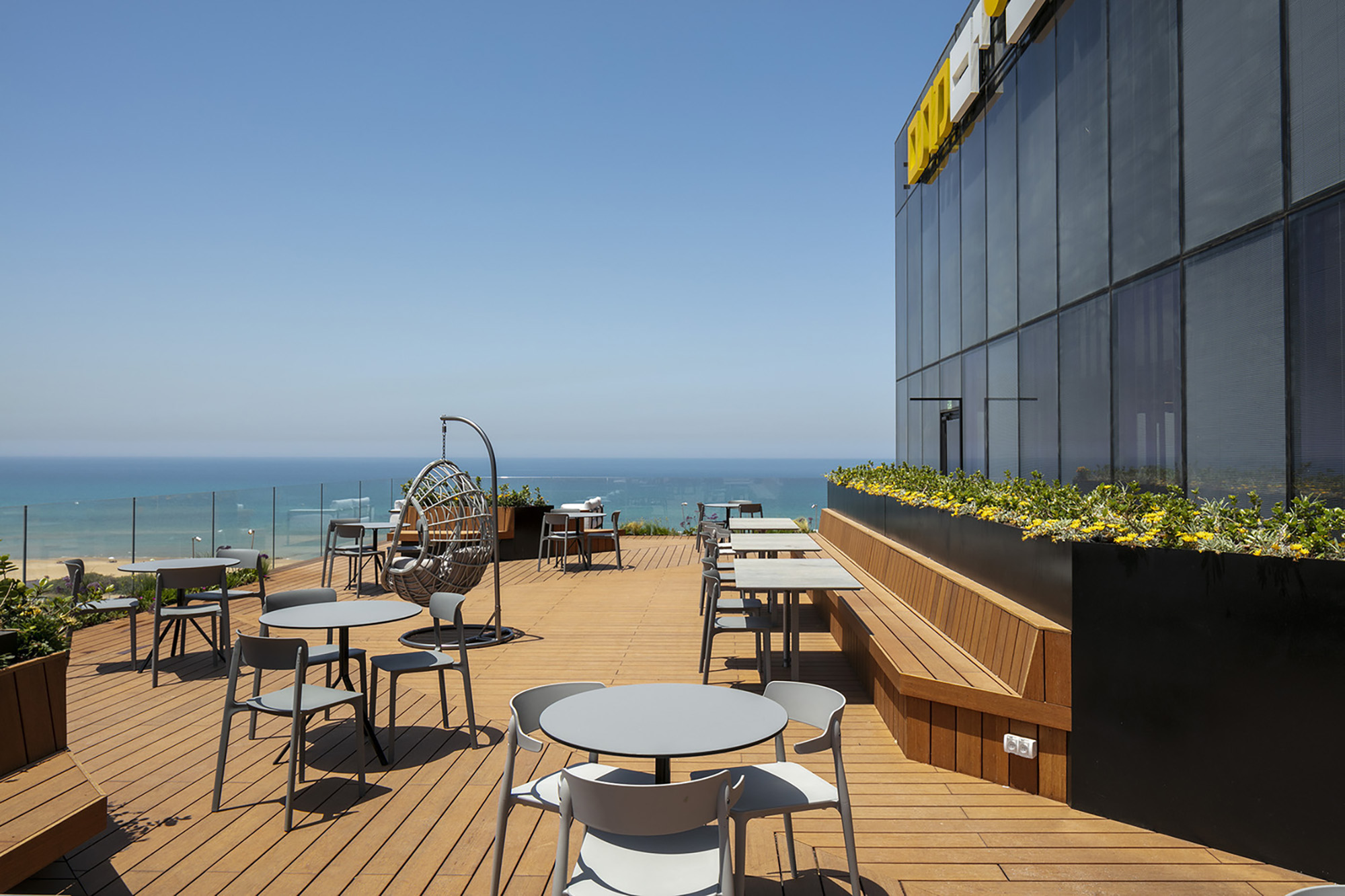 A modern rooftop terrace features sleek, minimalist furniture arranged on warm timber decking. Panoramic glass walls showcase a stunning ocean view, while vibrant planters filled with greenery enhance the inviting atmosphere. Contemporary design elements create a harmonious blend of comfort and elegance, perfect for outdoor dining or social gatherings.